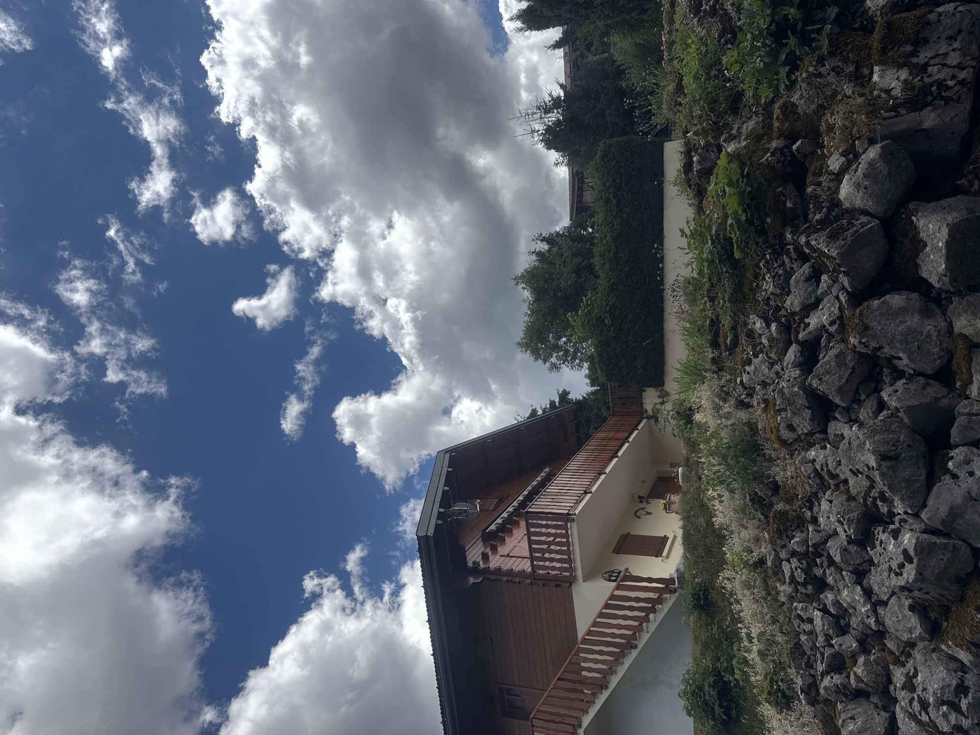 This is a medium-shot, eye-level, outdoor photograph taken on a sunny day with scattered clouds in Les Rousses, France. The image features a wooden chalet-style house with a red-brown exterior, a dark roof, and a balcony with a wooden railing. A white staircase with wooden handrails leads up to the balcony. The house is situated on a rocky slope covered with green vegetation and moss. In the background, there are dense green trees and hedges. The sky above is a vibrant blue with fluffy white and grey clouds. No people are visible in the image.