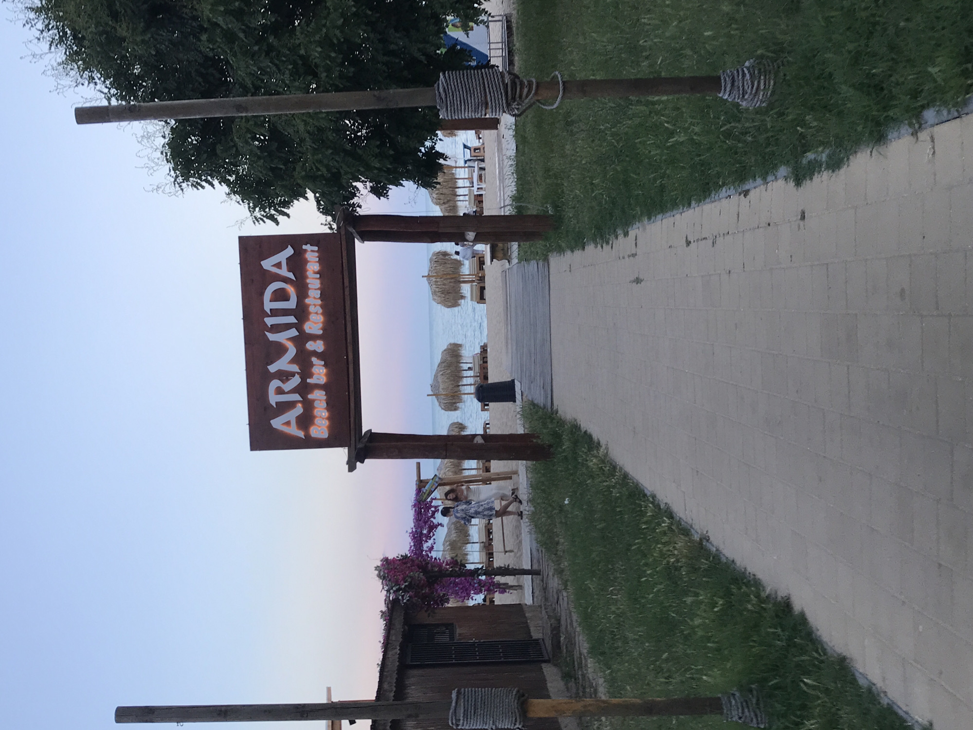 The image shows an entrance pathway leading to a beach bar and restaurant named "ARMIDA" in Varna, Bulgaria. The sign for the establishment is visible above a wooden archway. The pathway is made of light-colored paving stones and is flanked by grass. On the left side of the pathway, there is a wooden structure with a thatched roof, and some purple flowers are blooming on a trellis. Two women are walking on the beach in the background. The beach itself is visible with thatched umbrellas and lounge chairs. The sea is in the distance under a twilight sky. There are also two wooden posts with ropes wrapped around them, one on each side of the entrance.