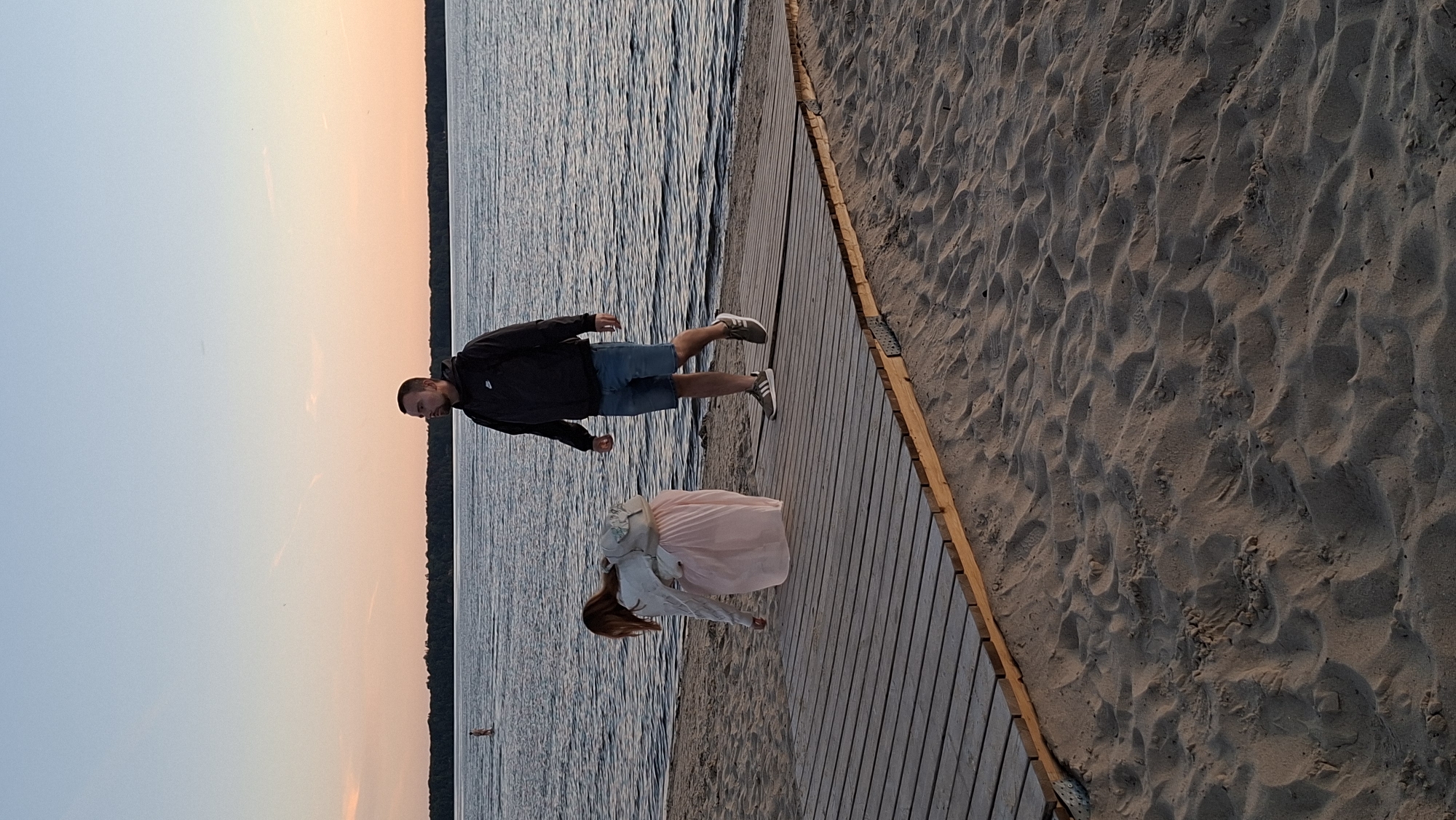 A man and a woman are on a wooden boardwalk at a sandy beach, possibly in Tallinn, Estonia, as the sun sets over a body of water. The man, wearing a dark hooded jacket and denim shorts, walks away from the viewer. The woman, in a light-colored jacket and a long, pale pink skirt, is bent over, appearing to pick something up from the boardwalk. She has a backpack slung over her shoulder. The sky displays a gradient of pale blue to peachy orange. In the background, a person is visible in the shallow water. The sand on the beach is disturbed by footprints and ripples.