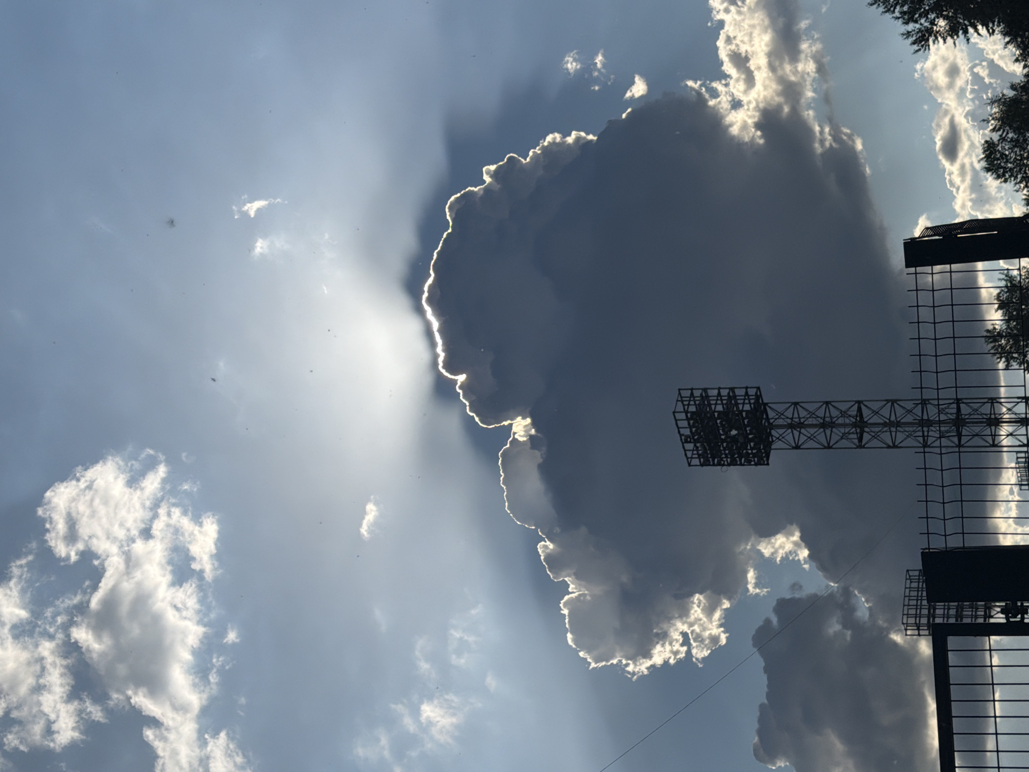 A low-angle, close-up shot captures a large, dark cloud dominating the sky, backlit by the sun. The sun's rays are piercing through the cloud, creating a halo effect and casting streaks of light across the blue sky. In the foreground, a tall, metal structure, likely a stadium light, is silhouetted against the dramatic cloud formation. A black chain-link fence runs across the bottom of the frame, with trees visible on the right edge. The scene suggests an outdoor, possibly urban setting, during daylight hours.