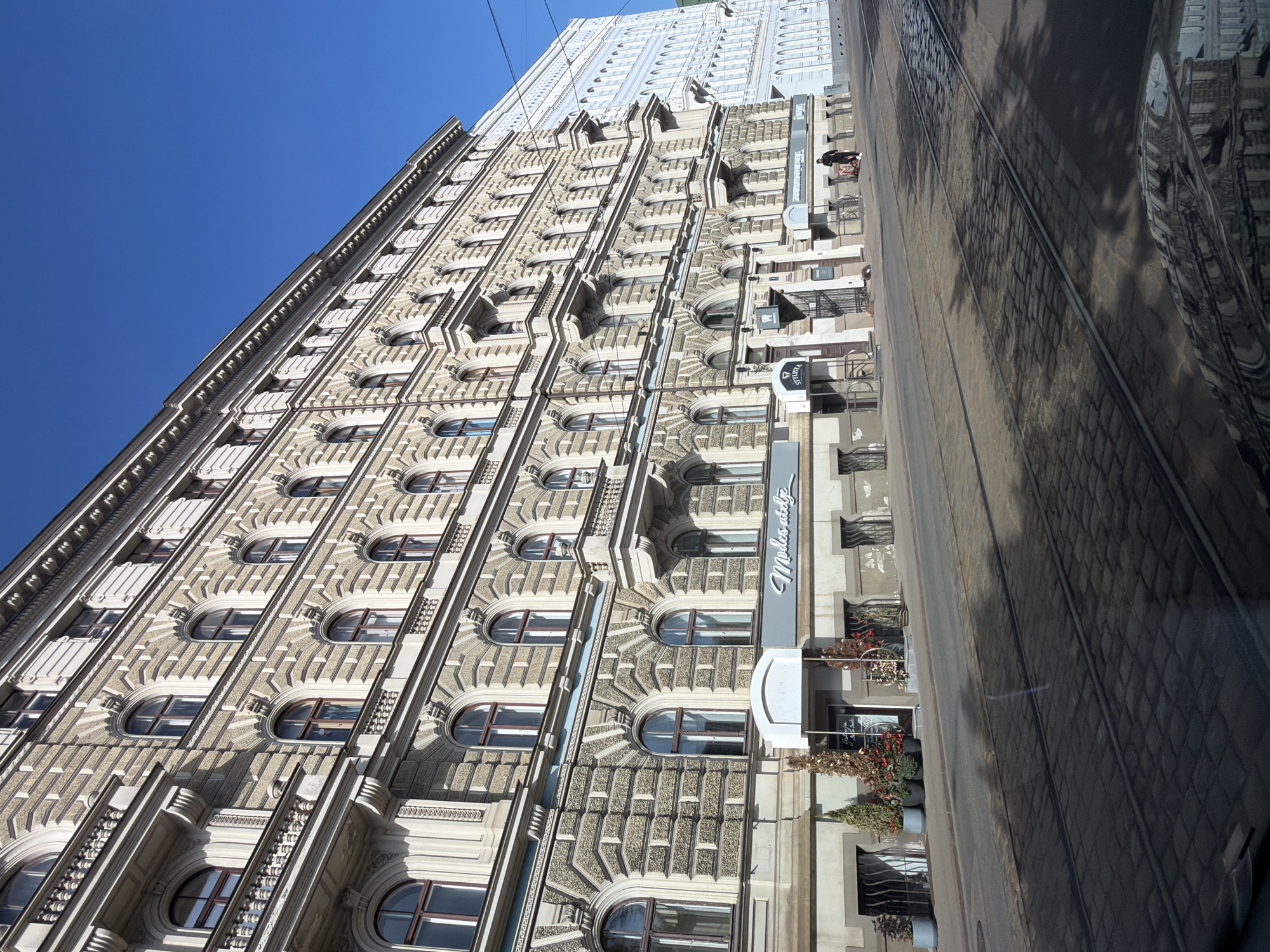 The image depicts a street view in Riga, Latvia, characterized by a grand, multi-story building with ornate architecture. The building is a light beige color with a textured facade, featuring numerous arched windows, balconies, and decorative molding. The street in the foreground is paved with cobblestones and has tram tracks running through it. Shadows from trees and possibly buildings are cast across the street.
Several businesses are visible on the ground floor of the building, including one with signage that appears to read "Modus atelje" and another labeled "Stylista." There are potted plants and trees outside the establishments, adding a touch of greenery. A person is seen walking on the sidewalk. The sky above is a clear, bright blue. The image captures a sunny day in what appears to be a historical district of Riga, with the architecture suggesting a late 19th or early 20th-century style.