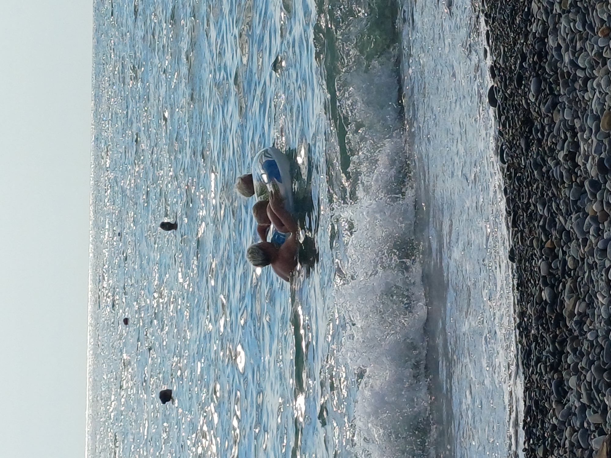 The image captures a serene beach scene along the coast of Sochi, Russia. In the foreground, a pebbled shoreline meets the gentle waves of the Black Sea. In the mid-ground, two individuals are enjoying the water. One person, with light-colored hair, is floating on a blue and white inflatable ring. They appear to be holding onto a smaller person, possibly a child, who is also partially submerged in the water. The sea is calm with shimmering sunlight reflecting off its surface. Several dark, round buoys are scattered across the water in the distance. The sky is a pale, hazy blue.