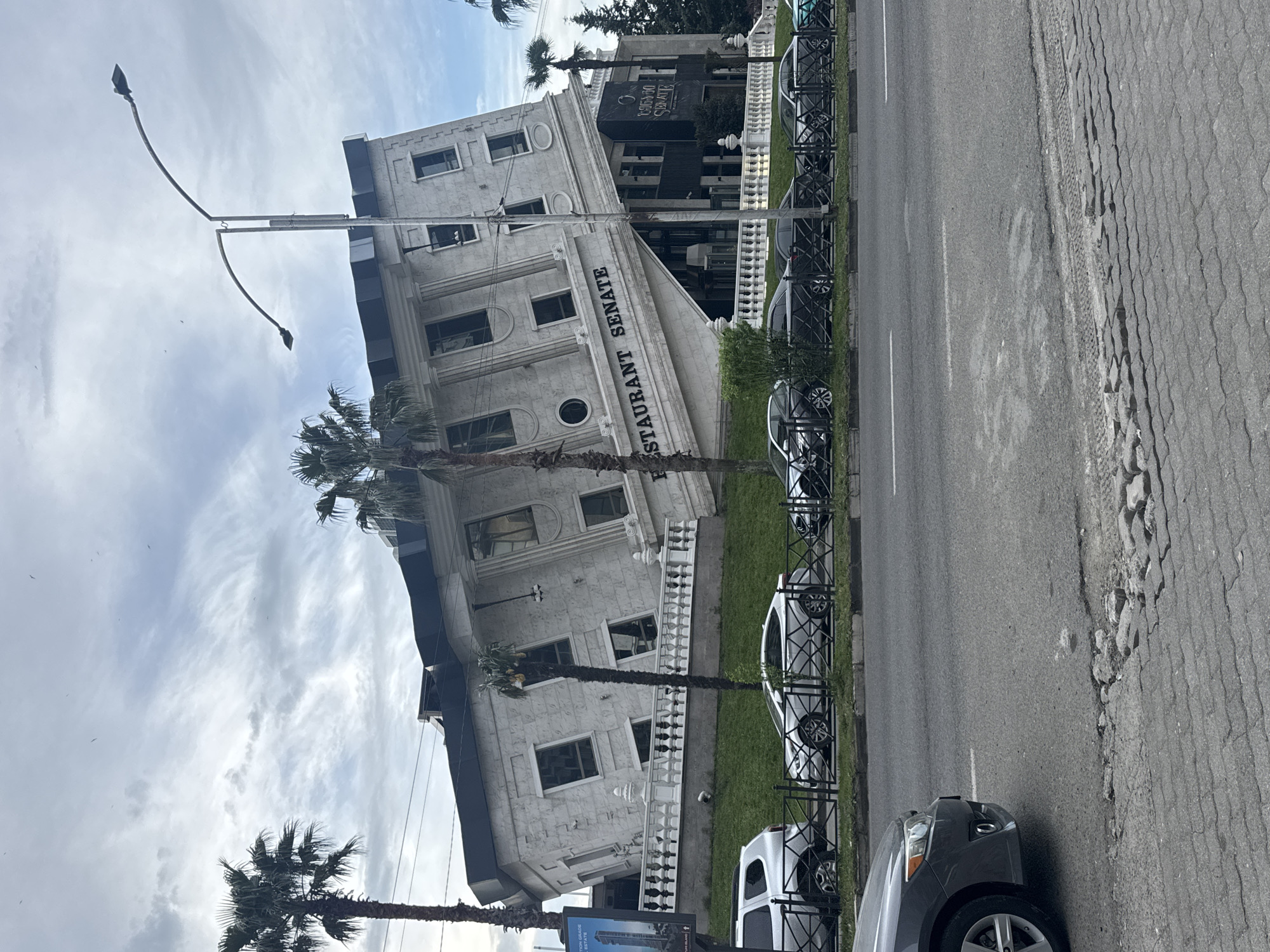 The image shows a building in Batumi, Georgia, identified as "Restaurant Senate". The building is light-colored, multi-story, and has a distinctive architectural style with decorative elements. A palm tree stands in front of the building. A street runs along the side of the building with cars parked along the curb. A sidewalk with damaged paving is visible. A lamppost is positioned near the building. The sky is cloudy with blue patches.