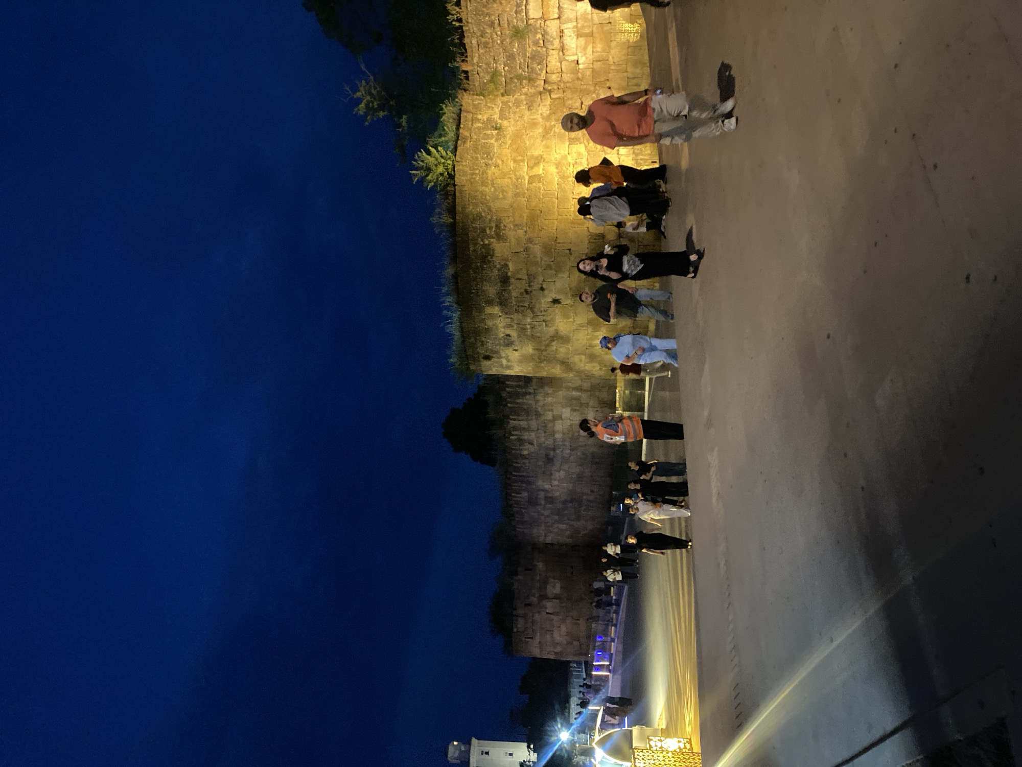 A night shot captures a wide stone wall, likely part of the ancient Derbent fortress in Russia, illuminated by warm yellow lights. The wall stretches across the upper portion of the frame, with a dark, deep blue sky above. A white tower or building with a light on top is visible in the far left background.
A group of people are walking on a wide concrete pathway in front of the wall. The pathway slopes slightly upwards from the foreground to the right. Several individuals are walking towards the right, while others are gathered in smaller groups. One person in an orange vest stands out, appearing to be a guide or security personnel, facing towards the left. The lighting creates distinct shadows on the pathway.