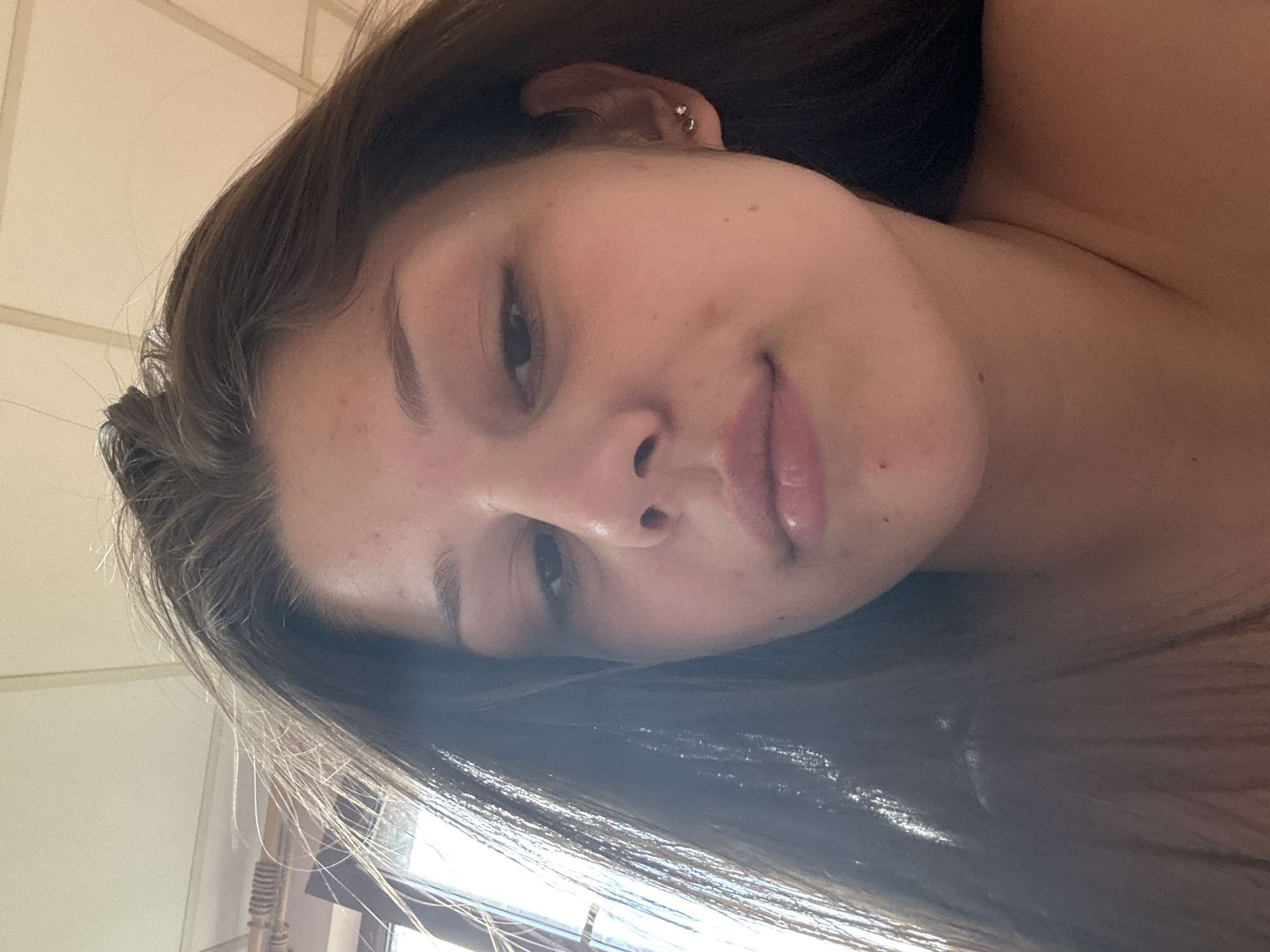 A close-up, low-angle, indoor selfie shows a young person with dark brown hair, fair skin, and light brown eyes. The person is looking slightly down and to the left, with a neutral expression. The lighting is bright, creating some glare on the hair. The background consists of a beige ceiling with recessed lighting and a window with blinds. There is a small, silver stud earring in the person's right ear. The person's shoulders and neck are visible.