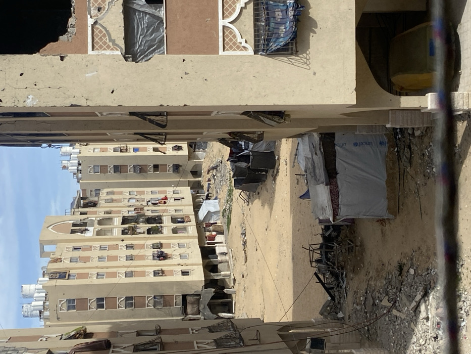 This image depicts a severely damaged residential area in Khan Yunis, Palestinian Territory, under clear daylight conditions. Multiple multi-story apartment buildings, constructed primarily of light-colored concrete or stucco with brown accents, frame an open, sandy ground area.
The building on the immediate right exhibits extensive structural damage, including a large, irregular cavity on an upper floor revealing interior space. Its facade is pockmarked with numerous impact points, consistent with bullet holes. Windows on this building are either shuttered, open, or covered with tarps and fabric, one prominently displaying a silvery-grey tarp, another a blue cloth behind a metal grille.
In the background, other similar apartment buildings stand, also showing signs of wear, disrepair, and numerous smaller impact marks on their facades. Many windows are open, covered, or have laundry hanging from them. White water tanks are visible on the rooftops of these structures.
The central ground area is an expanse of light-brown sand and dirt, scattered with debris and rubble, particularly in the immediate foreground left where twisted metal frames and concrete fragments are visible. Several makeshift shelters are present: a prominent one in the foreground right is constructed from assorted materials, including a white tarp section displaying the "UNICEF" logo. A row of at least four smaller, dark-colored, rudimentary shelters stretches across the mid-ground. Near the base of one of the background apartment buildings, a smaller white tent-like structure is visible.
One individual, dressed in dark clothing, is partially visible standing near the base of a background apartment building, adjacent to the small white tent. The overall scene suggests a degraded urban environment marked by extensive conflict damage and improvised living arrangements.