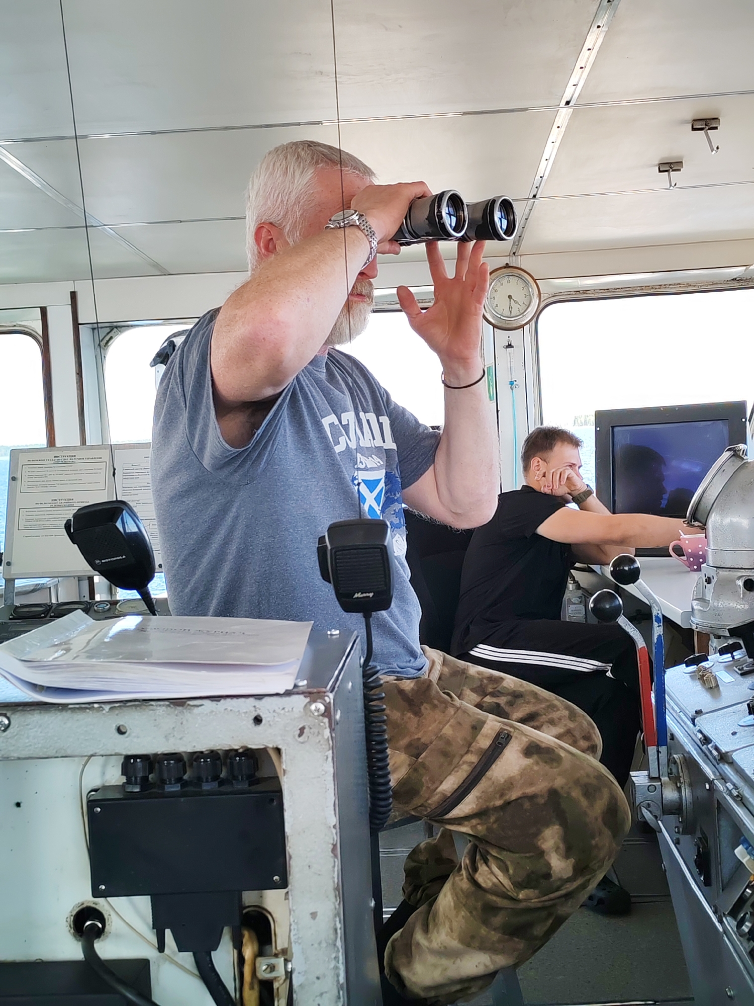 The image depicts the interior of a ship's bridge or control room, likely in Krivonogovskaya, Russia, during daytime under clear weather conditions. Two men are present within the cabin.
In the foreground, a man with a grey beard and hair is seated, actively using a pair of black binoculars, looking out through the large windows. He wears a grey t-shirt featuring a blue saltire flag design, suggesting "SCOTLAND" or a similar theme, and camouflage-patterned trousers. A silver watch adorns his left wrist. He is positioned behind a console equipped with two distinct microphones, one branded "Motorola" and the other "Mercury." Several documents are stacked on this console, and a box with visible cables and connectors is at its base.
To the right and further back, a second man with short brown hair is seated at another operational console. He wears a black t-shirt and black track pants with white stripes running down the sides. His posture is relaxed, with his right hand resting on his chin as he looks towards a monitor screen, which shows a faint reflection. A pink coffee cup with white polka dots sits on his console.
The cabin features a bright, paneled ceiling and large windows that illuminate the space. A traditional round clock with a white face and black hands is mounted on the wall between the windows. On a white board in the background, there is visible Cyrillic text, including "ИНСТРУКЦИИ" (Instructions), indicating operational guidelines or information. The overall scene suggests a functional setting aboard a vessel.