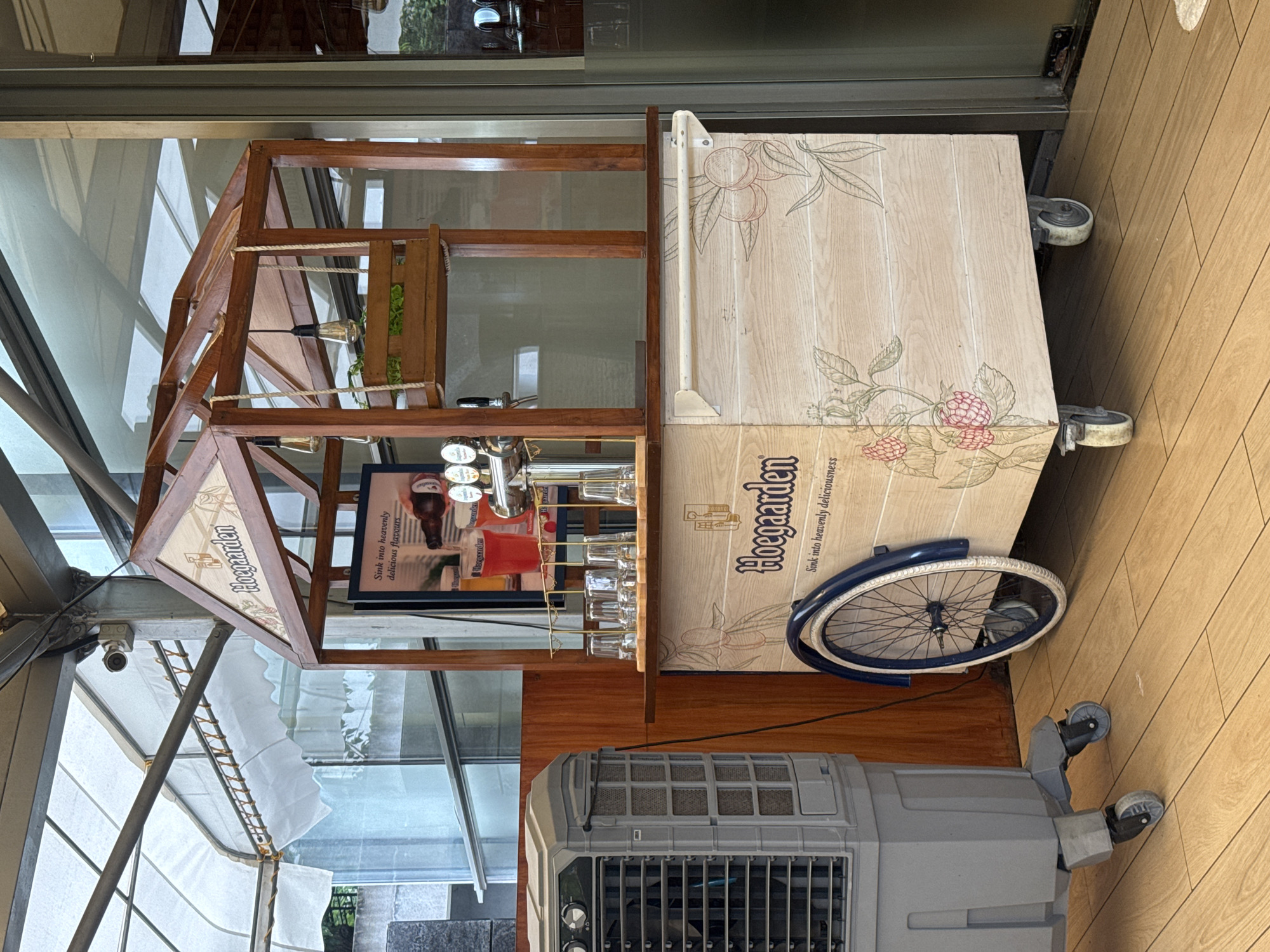 The image shows a portable beverage cart branded with "Hoegaarden" in an outdoor or semi-outdoor setting. The cart is made of wood and has a decorative wooden roof structure with hanging light bulbs. It is equipped with a beer tap system, glasses, and a sign that reads "Sink into heavenly delicious flavors." The cart's body is adorned with a light wood grain pattern and illustrations of fruits or berries. A bicycle wheel is attached to the side of the cart as a decorative element.
To the left of the cart is a large, gray evaporative cooler unit on wheels. The background reveals a building with large windows and a retractable awning, suggesting a commercial establishment such as a restaurant or bar. The floor is made of wooden planks. The overall scene appears to be a well-lit day.
Based on the context provided, this scene is located at The Flour Works, a restaurant in the city of Pune, India. The Flour Works is known for its rustic and charming ambiance, often featuring outdoor seating areas and decorative elements that complement a relaxed dining experience. The presence of a Hoegaarden branded cart is consistent with establishments that serve a variety of beverages, including popular international beers. The large evaporative cooler unit suggests a warm climate, which is typical of Pune for much of the year.