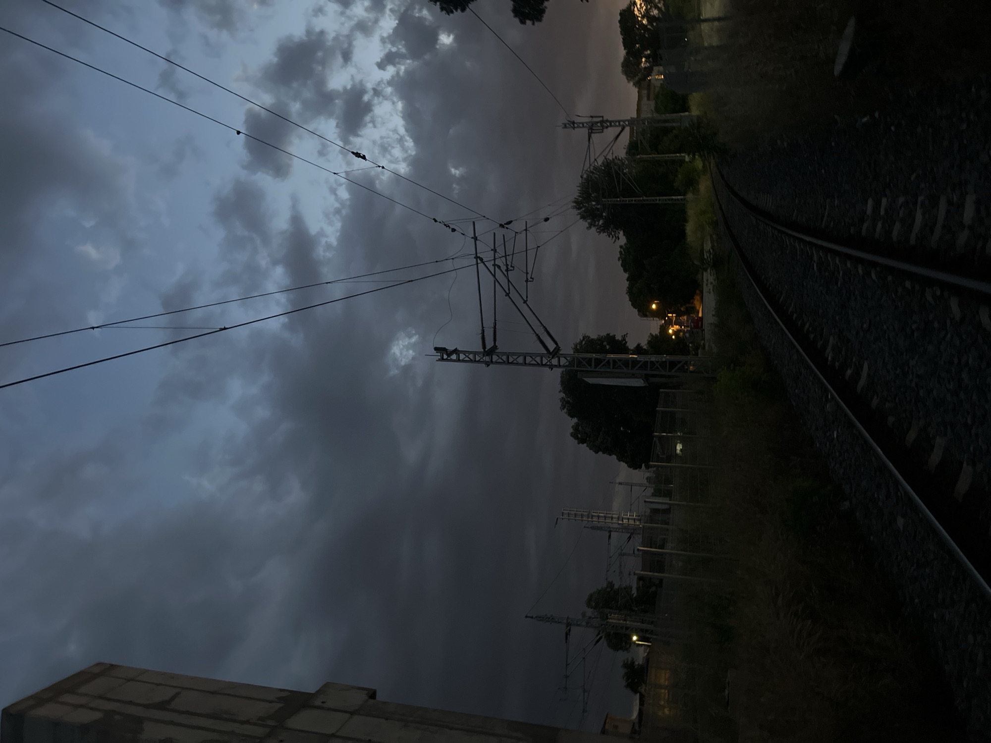 The image shows a railway track at dusk. Power lines and overhead gantries run above the tracks. Vegetation grows beside the tracks. A building is visible in the lower left of the frame. The sky is cloudy.