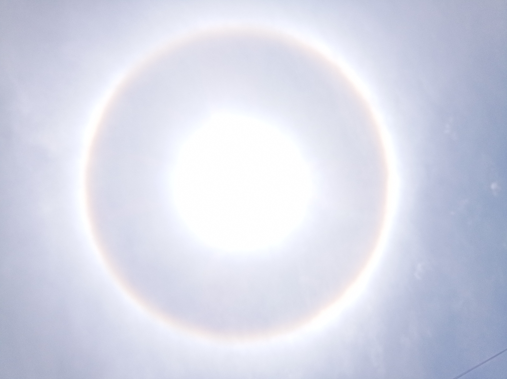 The image shows a 22° halo around the sun. The halo is a bright, circular ring of light surrounding the sun, with a faint rainbow coloring on its inner edge. The sky is light blue, with some indistinct clouds. The sun itself is a bright, white disc in the center of the halo. There is no evidence that the photo was taken in Porlamar, Venezuela. No people, subjects, objects, actions, or interactions can be identified beyond those previously listed.