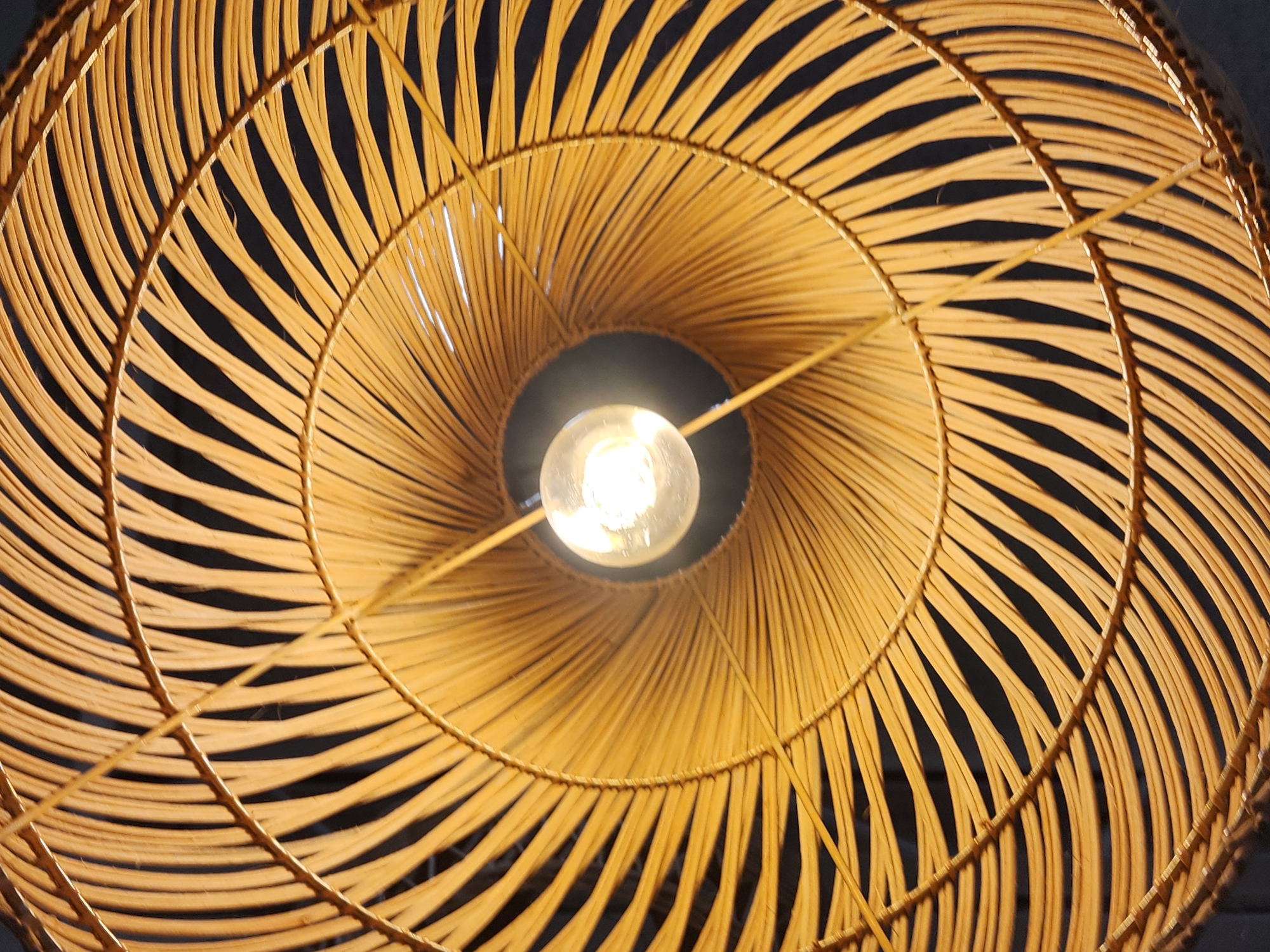 A low-angle, close-up shot captures the intricate details of a woven rattan lampshade. The lampshade, made of thin, natural-colored rattan strips, is arranged in a radial pattern, creating a fan-like effect. The strips are woven together to form concentric circles, with a central opening where a bare light bulb hangs. The light bulb is illuminated, casting a warm glow and creating shadows between the rattan strips, which appear dark against the bright background. The background itself is dark, likely indicating the interior of a room or a dark ceiling. There are no people or animals visible in the image. The scene appears to be an interior shot, possibly of a pendant light fixture in a home or commercial space. The image does not contain any information to determine its location in Maitland, United States of America.
