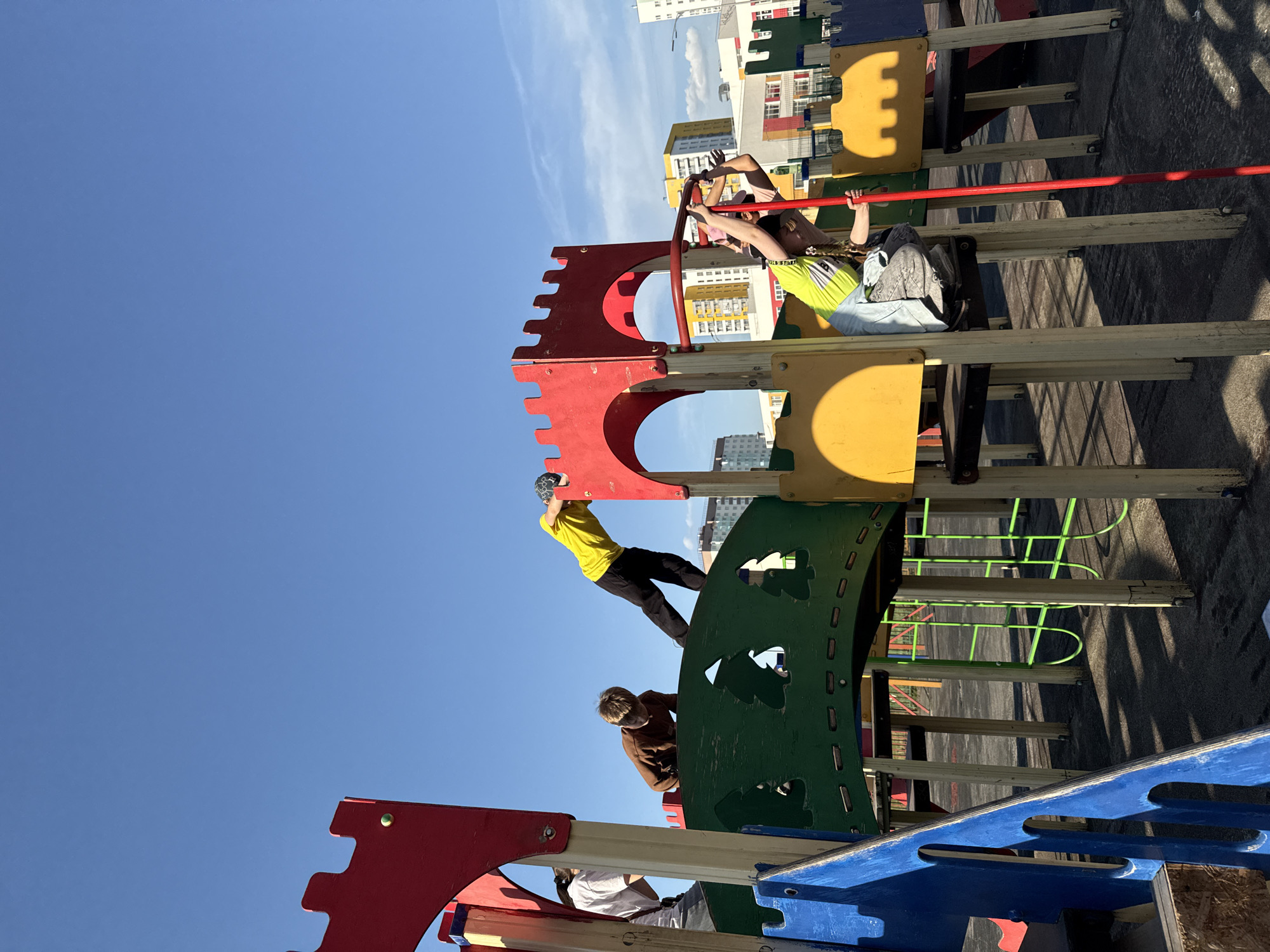 Three children are playing on a playground structure. The structure is multi-colored, with red, yellow, green, and blue elements. One child is hanging from a red bar at the top of the structure. Another child is climbing on a green curved wall. The third child is standing on a lower level of the structure. The sky is blue with some clouds. In the background, there are buildings, indicating a city location. The location is Tobolsk, Russia.