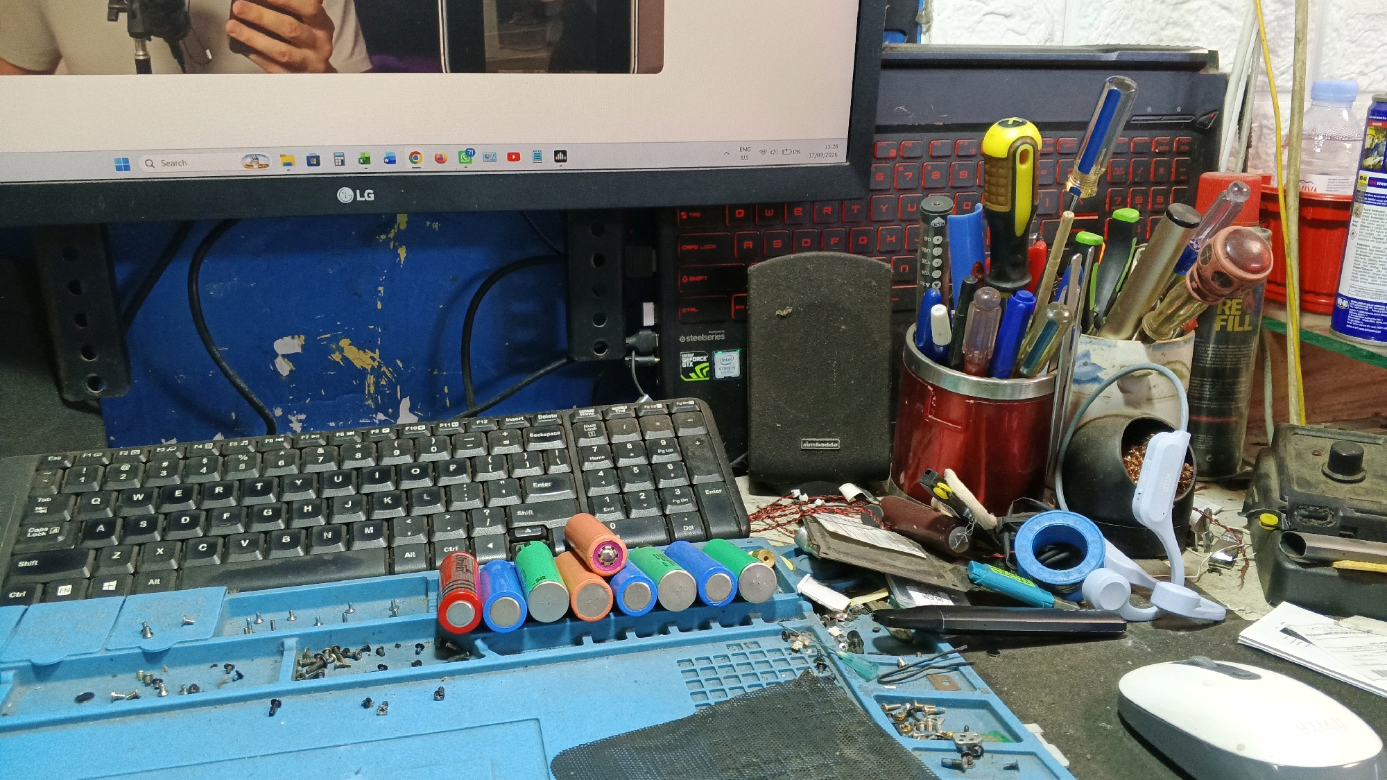 This image captures a cluttered workbench in what appears to be a repair or electronics workshop. The scene is dominated by electronic components and tools. In the foreground, a blue mat with compartments holds various small screws and electronic parts. A row of colorful cylindrical batteries, including red, blue, green, and orange, are laid out on this mat. Behind them, a black keyboard is visible, with its keys showing wear.
To the right of the keyboard, a collection of tools is organized in containers. These include screwdrivers with various colored handles (yellow, blue, green) and other small repair implements. There is also a red metal cup holding more writing utensils and small tools. Further back, a black speaker sits on top of a red-lit keyboard, suggesting a computer setup.
Various other items populate the scene, including a white computer mouse, a roll of blue tape, a black electronic device with a lens, and several bottles, one of which is white and red with text, possibly containing cleaning fluid or lubricant. The background features a blue surface with yellow paint splatters, and a portion of a computer monitor is visible at the top of the image, showing a person's hands holding something, possibly a phone or tool.
The overall impression is of a functional, albeit messy, workspace dedicated to electronic repairs. The presence of tools, components, and a computer setup indicates a hands-on, technical environment. The lighting is somewhat dim, suggesting an indoor setting, possibly a home workshop or a small repair shop. The image does not provide any specific indicators of location within Subah, Indonesia, beyond the general context of a workshop.