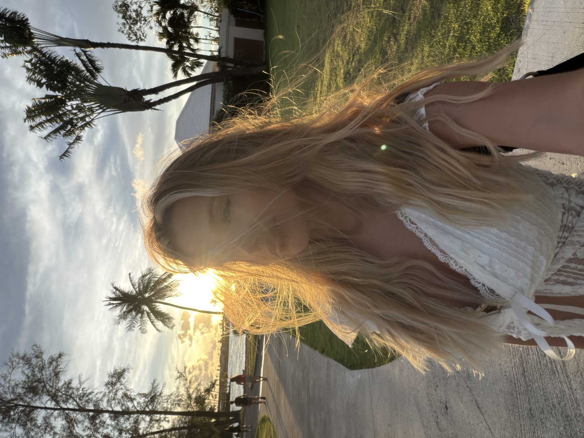 This is a vertical, low-angle, close-up selfie of a young woman with long, blonde hair against a backdrop of palm trees and a sunset. The woman is wearing a white lace top. Her hair is blowing in the wind and partially obscures her face. The sun is low in the sky, casting a warm, golden light and creating a lens flare. The background shows a path, a body of water, more palm trees, and a building. The overall mood is peaceful and idyllic, evoking a sense of a tropical vacation. The time of day is sunset. The weather appears to be clear and warm. The location is likely a beach or a resort area in a tropical climate, suggested by the palm trees and the warm lighting. There is no visible text in the image.