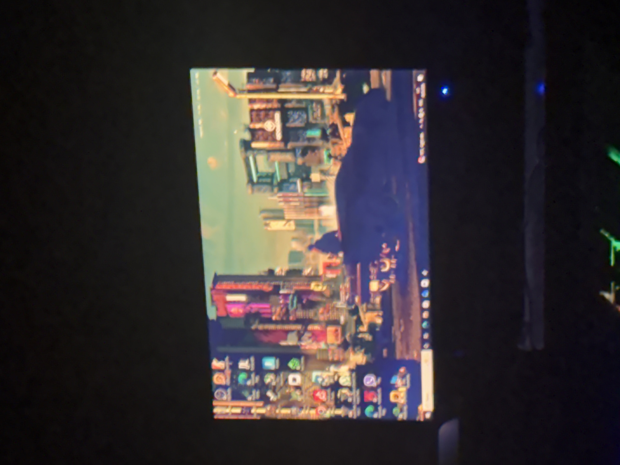The image shows a computer monitor in a dark room. The screen displays a desktop background with a city skyline. The taskbar at the bottom of the screen shows a variety of application icons. There are two blue lights visible on the monitor frame. A portion of a keyboard with green illumination is visible on the right side of the image. There is no indication of Yerevan, Armenia.