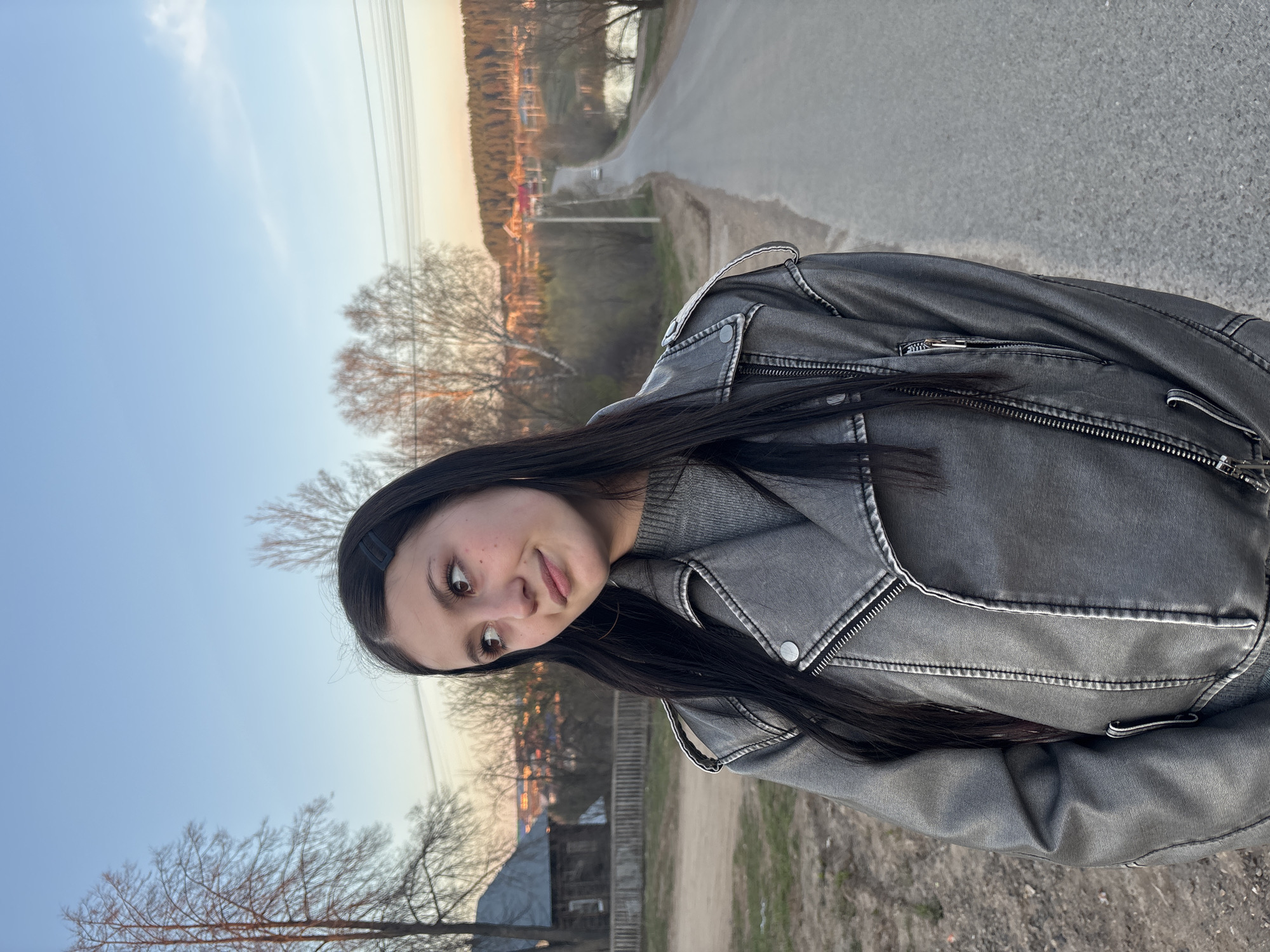 A young woman with long, dark hair and a black hair clip stands on the side of a road in Фоки, Russia. She is wearing a grey leather jacket and a grey sweater. She looks to her left with a slight smile. The road is asphalt and runs past a field of dry grass and trees. In the background, there are buildings and more trees. The sky is a pale blue near the horizon, transitioning to a lighter blue higher up. It appears to be dusk.