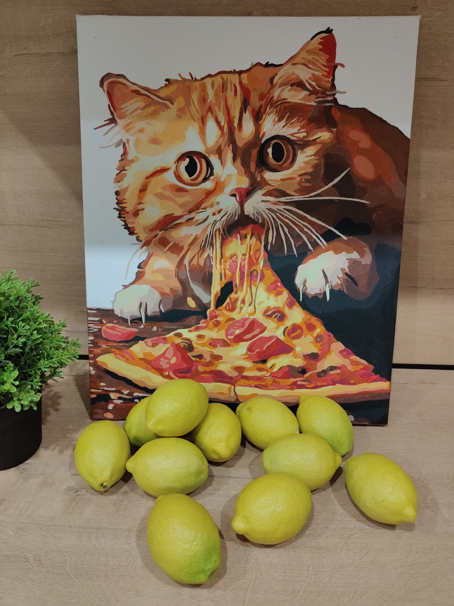 A staged composition on a wooden surface featuring a printed canvas of a cat eating a slice of pizza behind a pile of lemons and a potted plant. The canvas depicts an orange tabby cat with a determined expression, its mouth latched onto a large slice of pizza, with strings of cheese dangling from its mouth. In front of the canvas, a collection of approximately 11 lemons are arranged, some clustered together and others scattered. To the left of the canvas, a small potted plant with dense green foliage sits in a dark pot. The wooden surface provides a neutral backdrop for the arrangement. No people or specific location are evident.