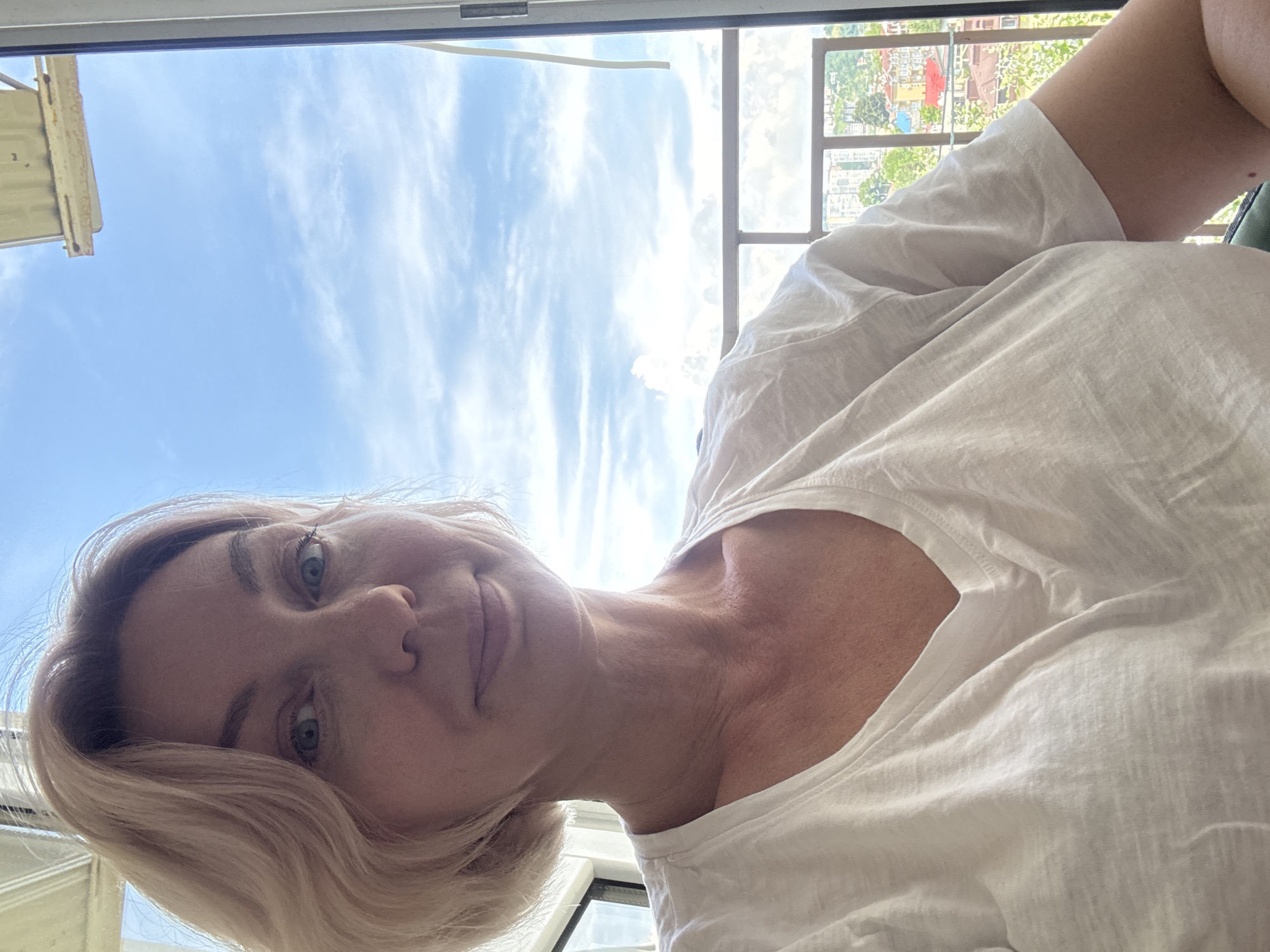 A woman with blonde, chin-length hair and blue eyes looks directly at the camera. She is wearing a white V-neck t-shirt. The background shows a bright blue sky with scattered clouds, a railing, and a cityscape that includes buildings with red and yellow roofs, and lush green trees. The scene appears to be from a balcony or window overlooking a city. This scene is not located in Sochi, Russia.