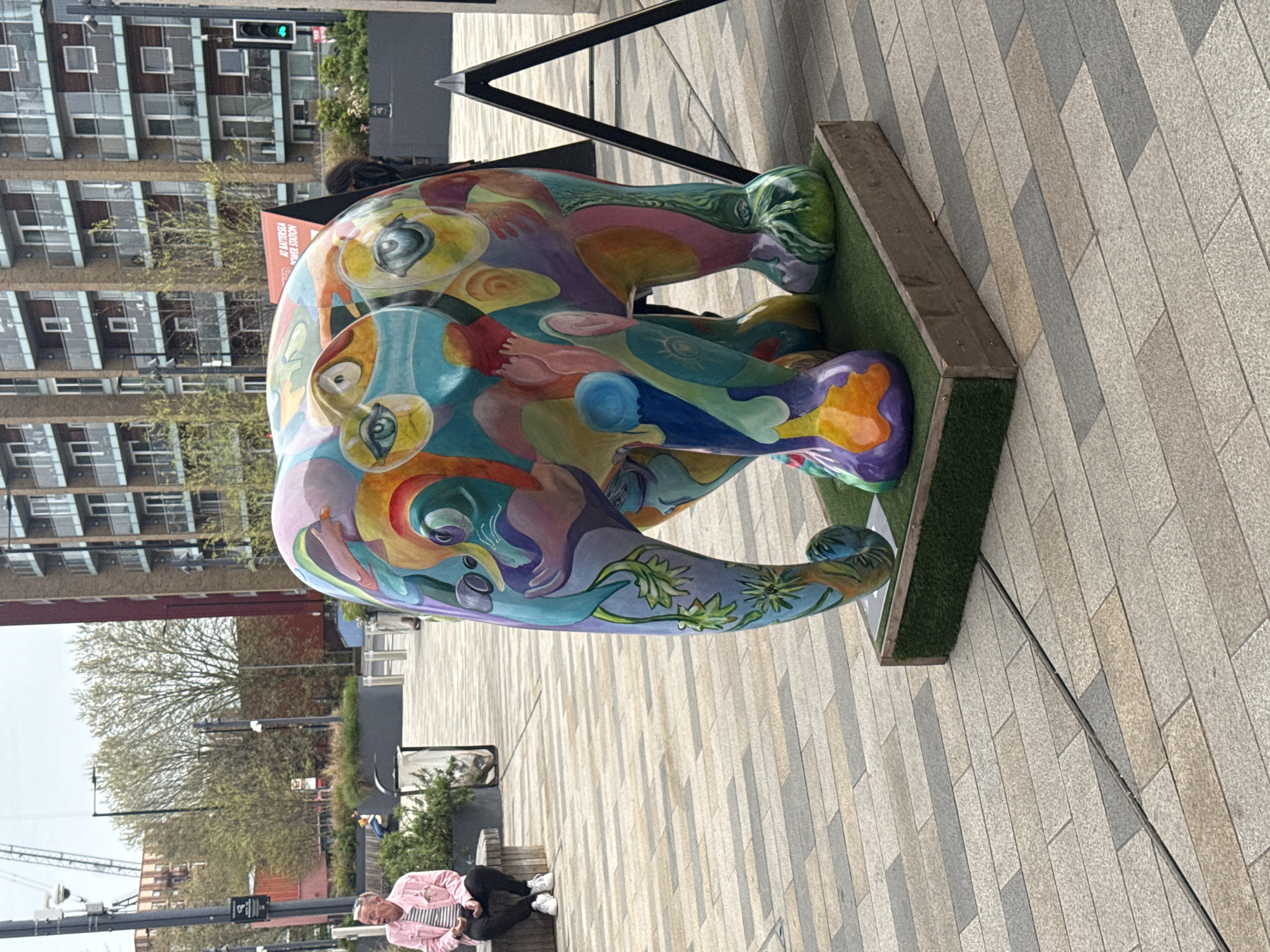 The image shows a brightly colored elephant sculpture on a raised platform with artificial turf. The sculpture features a variety of colors and patterns, including what appears to be depictions of faces and floral designs. A metal frame structure is visible behind the sculpture.
In the background, there is a modern building with numerous windows, as well as a street scene with trees and streetlights. Two people are sitting on a low wall in the foreground. One is wearing a pink shirt.
The location is London, United Kingdom. The presence of the elephant sculpture suggests it may be part of an art installation or public art display. The modern architecture in the background indicates a contemporary urban environment.