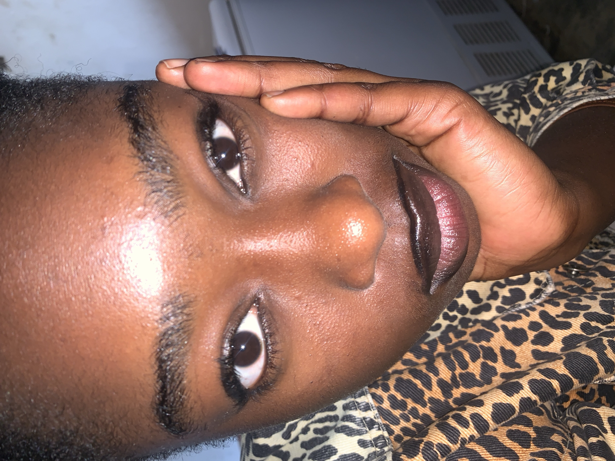 A close-up, vertically oriented, low-angle shot captures the face of a young Black person. Their eyes are open, with one eye fully visible and the other partially obscured by a hand resting on their forehead. The subject is wearing dark lipstick and their skin appears smooth and dewy. Their eyebrows are dark and well-defined. The background is blurred, showing hints of a white surface with horizontal ridges and a garment with a leopard print pattern. The lighting suggests an indoor setting, possibly with artificial light. The overall mood is contemplative or perhaps slightly pensive. There is no visible text.