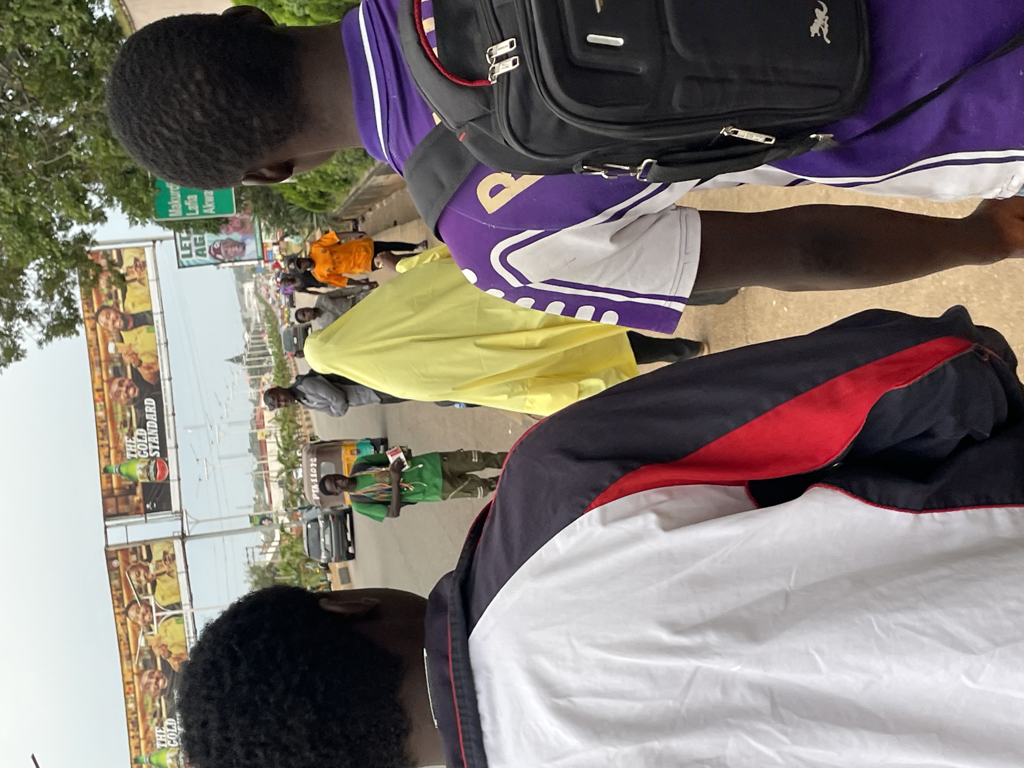 This is a medium shot of two young men walking in Jos, Nigeria, with their backs to the camera. They are on a paved sidewalk next to a road. The man on the right wears a purple shirt with white trim and a black backpack. The man on the left wears a white, red, and black jacket. In the background, there is a billboard advertising "The Gold Standard" with images of men holding bottles. There are vehicles, a yellow auto-rickshaw, and other pedestrians. The environment appears to be urban with some greenery.