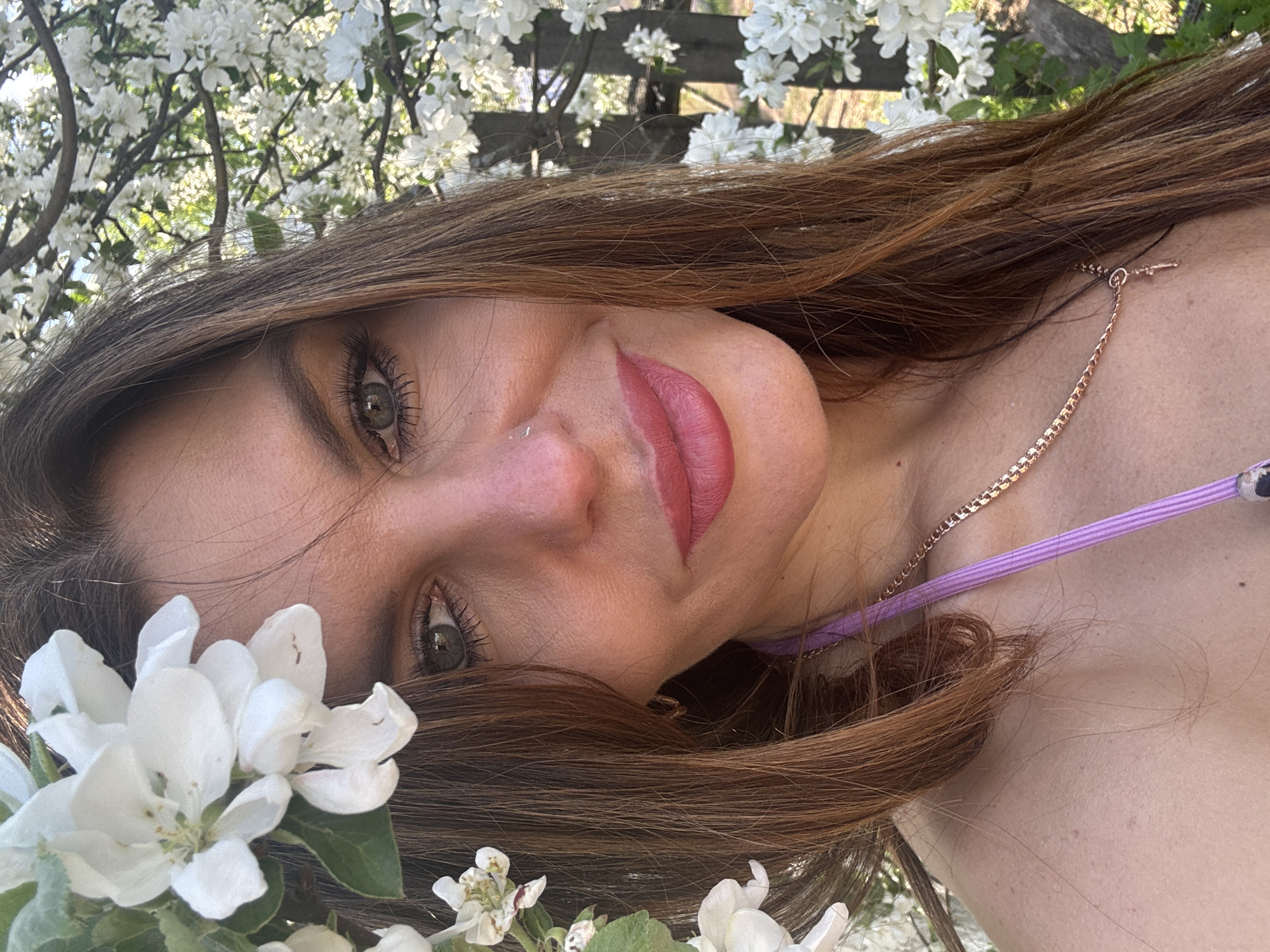 A woman with light skin, brown hair, and green eyes is positioned among white flowers. She wears a light purple strap and a gold chain necklace. The background includes more white flowers and a wooden structure. It is unknown if the location is Dubovaya, Russia.