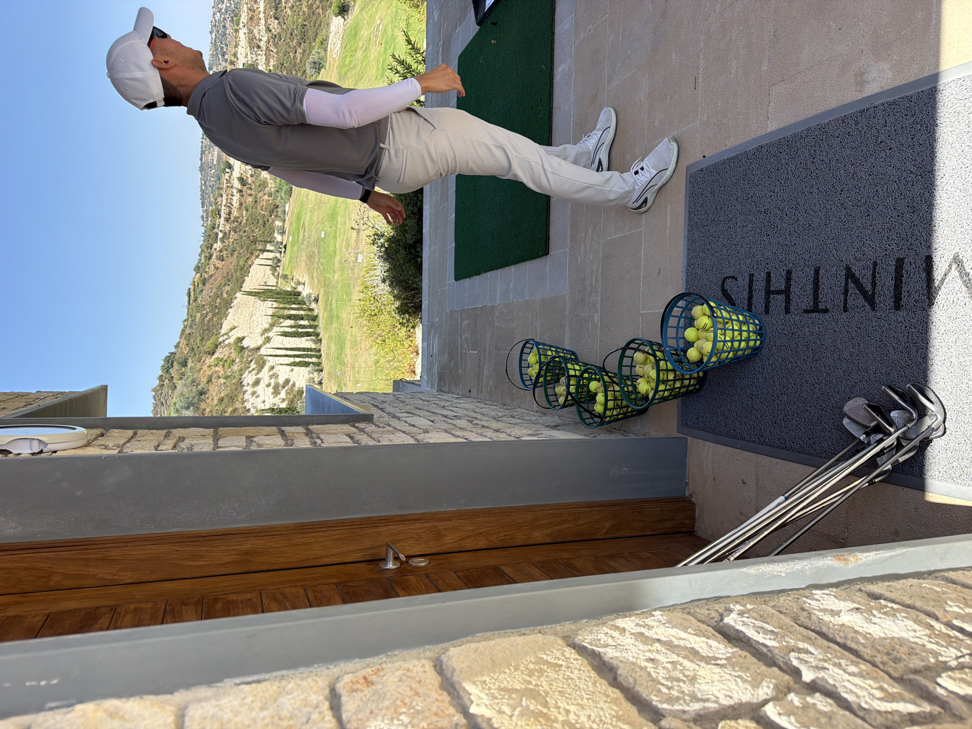 A man, wearing a white baseball cap, grey polo shirt, and light-colored pants with white arm sleeves, stands on a paved area at a golf course in Tsada, Cyprus. He is facing away from the camera, looking towards a golf course with a hilly landscape under a clear blue sky. In front of him, lined up on a grey mat, are five green baskets overflowing with yellow golf balls. To the left of the baskets and mat, leaning against a stone wall and wooden door, is a set of golf clubs. The setting appears to be a driving range.