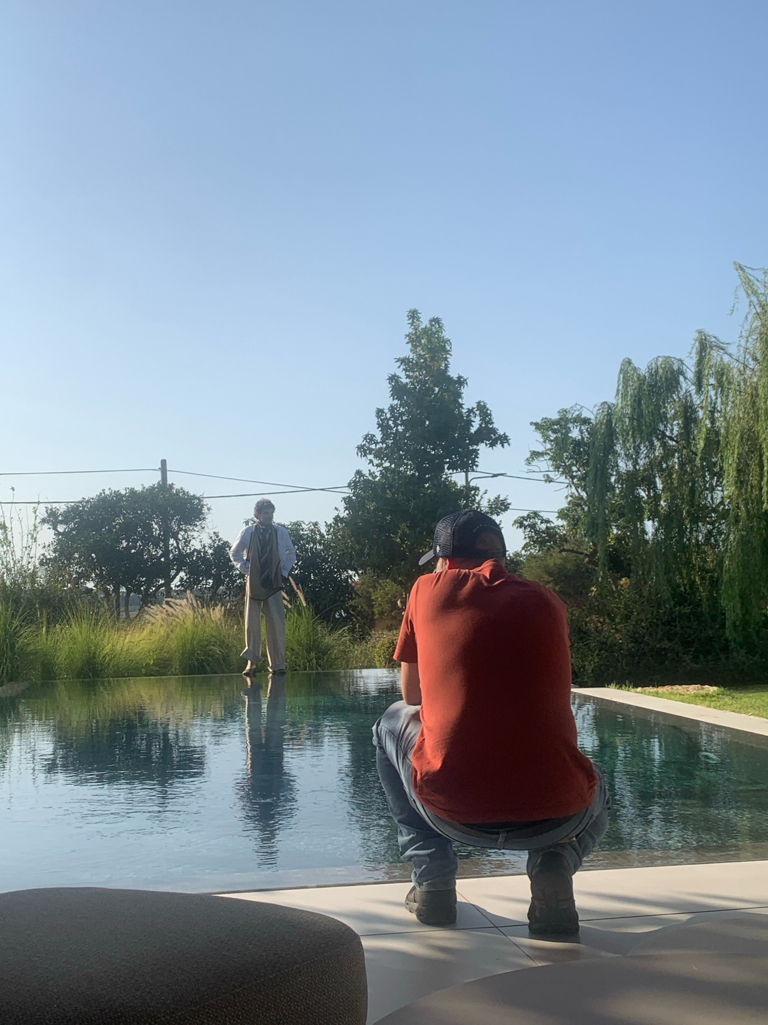 Two men are positioned in a backyard setting in Kfar Monash, Israel, near a rectangular swimming pool. One man is standing in the pool, facing away from the camera, dressed in a white collared shirt, khaki pants, and a vest with a scarf around his neck. The other man is in the foreground, facing away from the camera, crouched down at the edge of the pool, wearing an orange t-shirt, blue jeans, and a baseball cap. The pool reflects the sky and the trees in the background. The environment includes lush green vegetation, trees, and a clear blue sky.