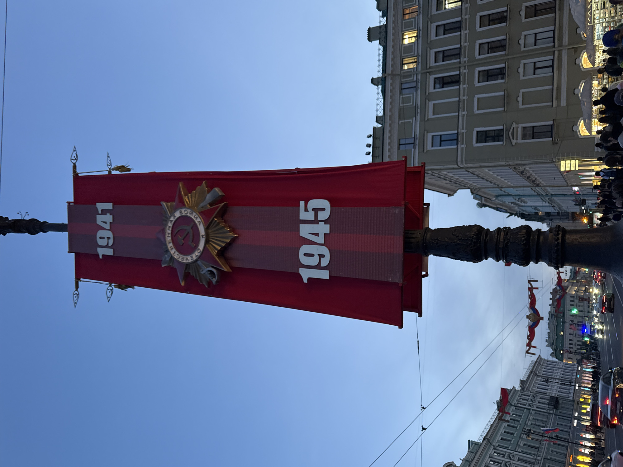 The image captures a street scene in Saint Petersburg, Russia, with a prominent red banner displayed on a lamppost. The banner features the years "1941" and "1945" in white numerals, separated by a Soviet-era military decoration on a ribbon. Buildings with multiple windows line the street, with people visible on the sidewalks. Banners strung between buildings are also visible. Cars and streetlights are present along the street. The sky is a clear blue.