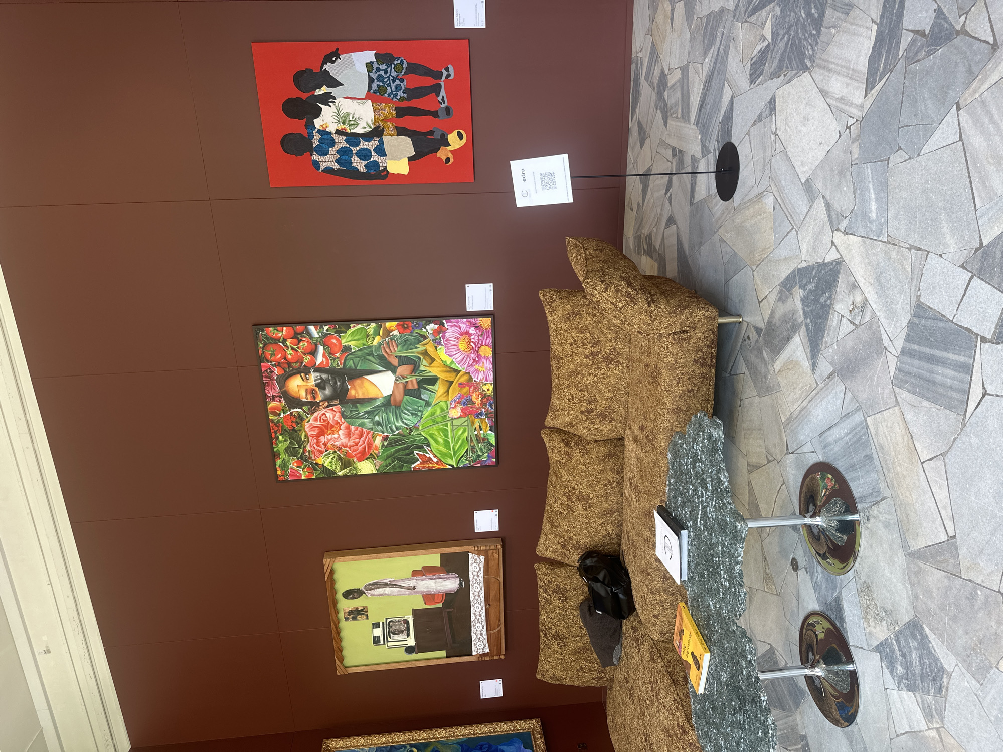 The image shows an art exhibition space. A mustard-colored sectional sofa with a textured fabric is positioned in the foreground, with a black bag and a gray sweater draped over it. In front of the sofa is a two-tiered coffee table with a textured, greenish-gray top and chrome bases. Two books are placed on the coffee table. The walls are painted a deep red. On the wall are three artworks. To the left, a framed painting shows a person in a white robe standing in a room with a television. In the center, a large painting features a person in a green jacket surrounded by vibrant flowers and tomatoes. To the right, a painting on a red background depicts the silhouettes of three children. A white sign with a QR code and the word "cedra" is displayed on a black stand near the right side of the sofa. The floor is covered in irregularly shaped gray and white stone tiles. The overall setting appears to be a contemporary art gallery.