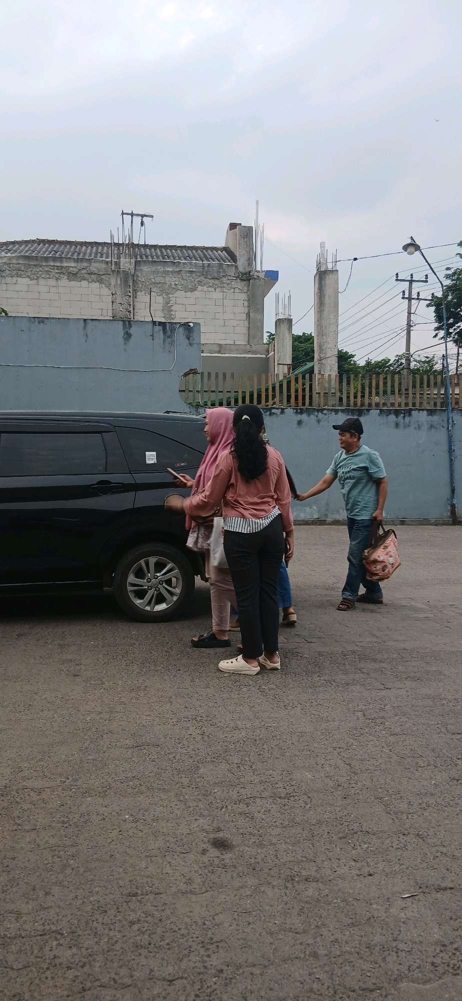 The image depicts an outdoor scene in Metro, Indonesia, possibly a parking lot or street. A black SUV is parked on the left. Three individuals are visible near the vehicle. On the left, a woman wearing a pink hijab and a pink long-sleeved shirt stands next to the SUV, holding a mobile phone. To her right, another woman in a pink long-sleeved shirt and black pants stands facing away from the camera. A man in a light blue t-shirt, dark baseball cap, and blue jeans stands to the right of the women, facing them and holding a patterned bag. He appears to be interacting with the women, possibly handing them the bag or receiving it. The environment includes a grey concrete wall in the background, a wooden fence, and utility poles with wires. The ground is paved with asphalt. The sky is overcast.