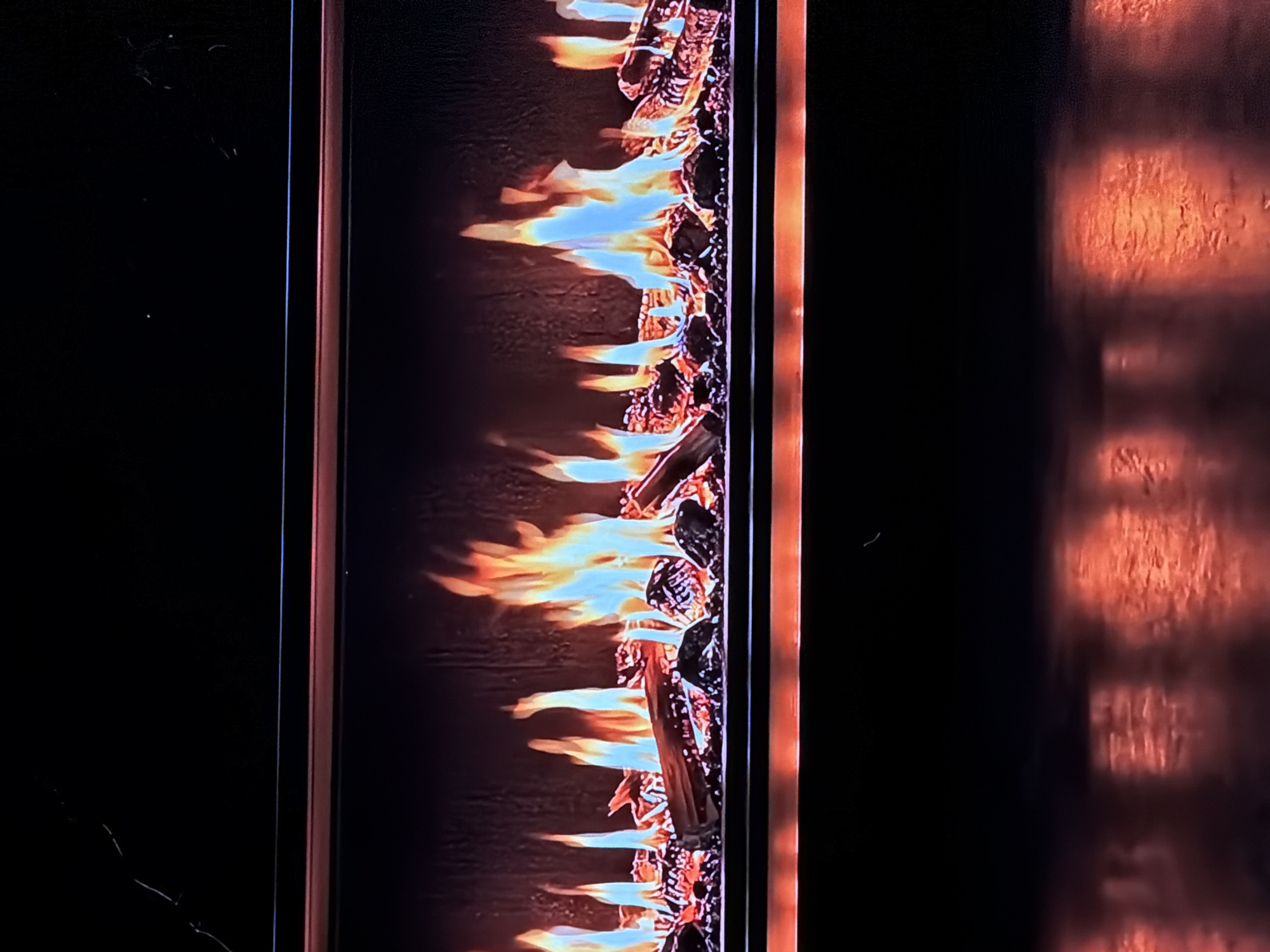 The media file captures a close-up, vertical view of a modern fireplace or fire feature, likely an electric or gas unit, situated in an indoor setting. The primary subject is a lively display of artificial flames rising from a bed of dark, textured, simulated logs or coals. The flames exhibit a spectrum of colors, transitioning from bright blue at their base to vibrant yellow and orange, creating a realistic and dynamic fire effect.
The fire feature is framed by a dark border on its left. To the right of this border, a textured, reddish-orange surface, possibly a wall or part of the fireplace unit, is softly illuminated by the glow of the flames, showing subtle patterns. The surrounding areas on both sides are predominantly black, indicating a dimly lit environment, characteristic of an evening or night setting. Minor dust or surface imperfections are visible on these dark surfaces. No people are present, and no text is visible. The scene evokes a sense of warmth and ambiance, originating from Sofiivska Borshchahivka, Ukraine.