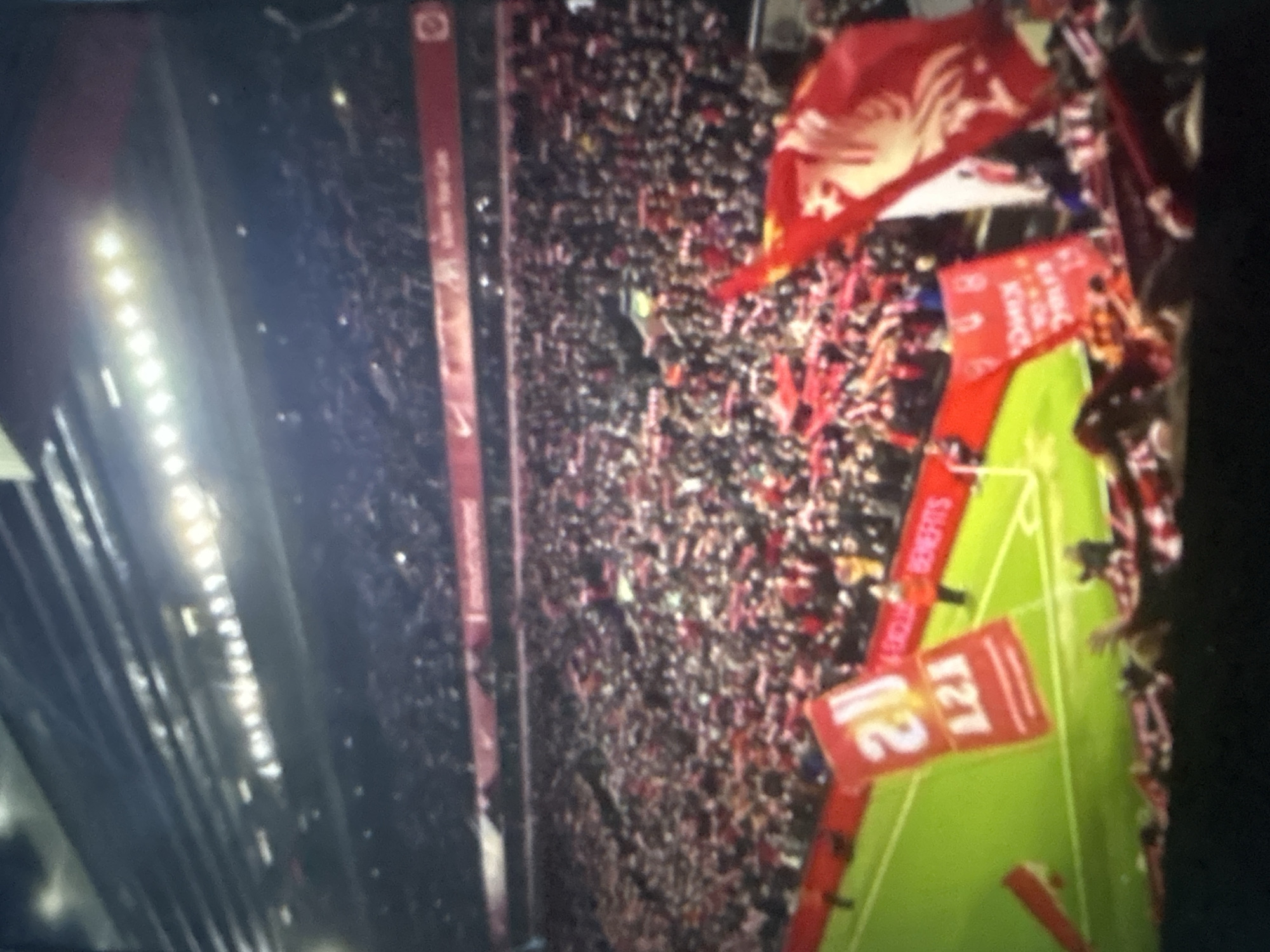 The image captures a vibrant night scene inside a densely packed football stadium, viewed from an elevated position. The stands are filled with a large crowd of spectators, predominantly dressed in red, indicative of strong fan support. Many individuals are waving red flags, some clearly displaying the white Liver bird emblem, strongly associated with Liverpool Football Club. Bright stadium floodlights illuminate the scene from the left, casting a glow over the upper sections of the stadium.
A green football pitch is visible in the foreground, featuring white line markings for a goal area. On or near the pitch, red banners or tarpaulins are displayed. One banner partially shows the word "BENEFITS", while another prominently displays the numbers "121" and "21". A vertical red panel along the stadium wall or advertising hoarding on the left side features the "NIKE" logo and some indistinct text above it. The atmosphere appears to be one of excitement and high energy, typical of a major sporting event. The time of day is clearly night due to the prominent artificial lighting.