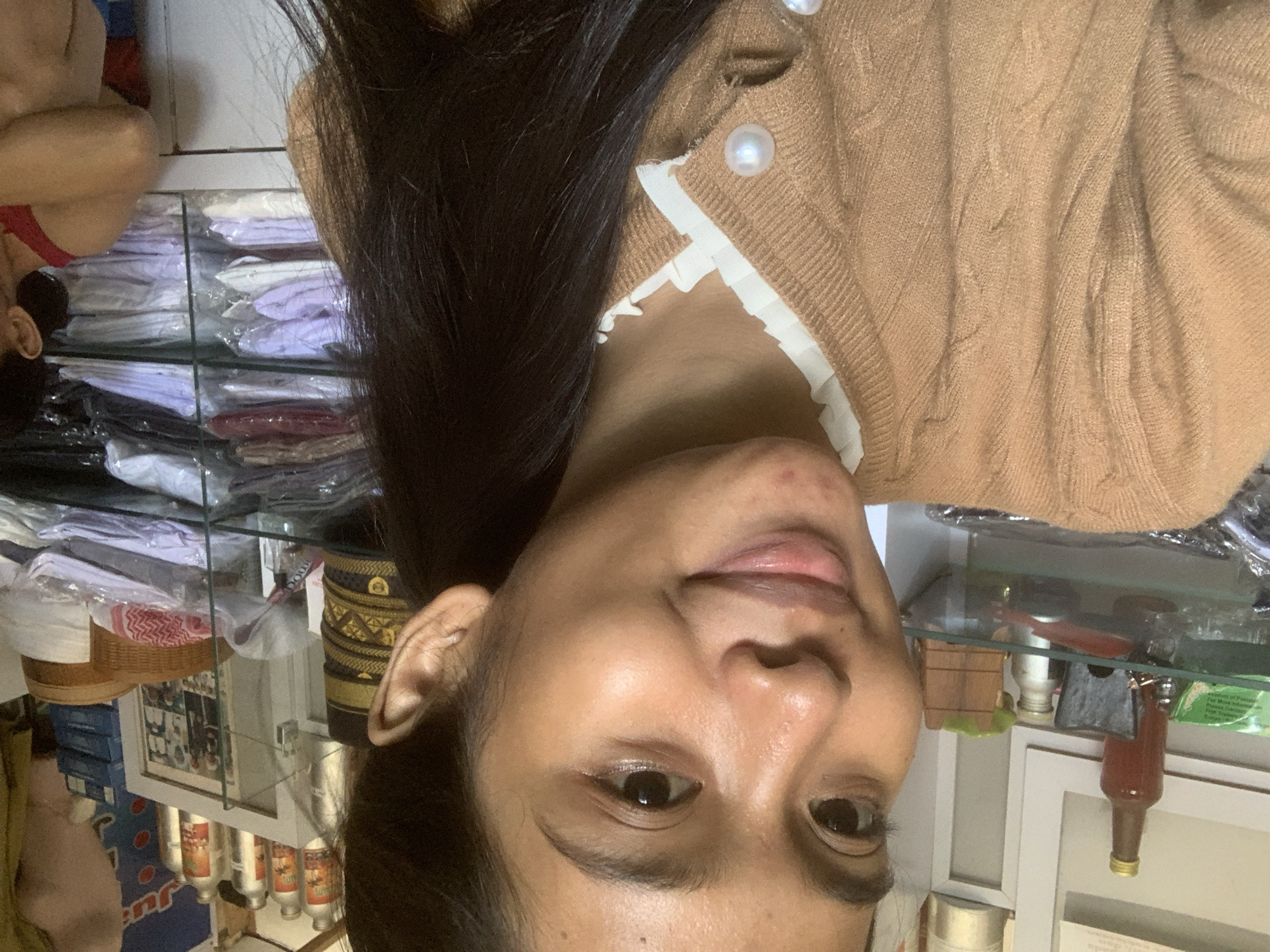 The image shows a close-up of a young woman, likely in her late teens or early twenties, with dark hair and brown eyes. She is wearing a brown knitted cardigan over a white ruffled-collar shirt. The cardigan is fastened with pearl-like buttons. Her skin appears clear with some subtle makeup. Her expression is a slight smile.
The background suggests an indoor environment, possibly a shop. Shelves are visible behind her, filled with various items. On the left, there is a shelf with bottles, one of which appears to be a small, dark glass bottle with a gold cap. Further back, on another shelf, there are stacks of folded garments wrapped in clear plastic, suggesting they are for sale. There are also some baskets and framed pictures visible in the background.
The presence of the clothing racks and display shelves strongly indicates a retail setting. The overall impression is that of a casual selfie taken within a shop environment. The mention of Sungai Meriam, Indonesia, places the scene within that specific geographical location.