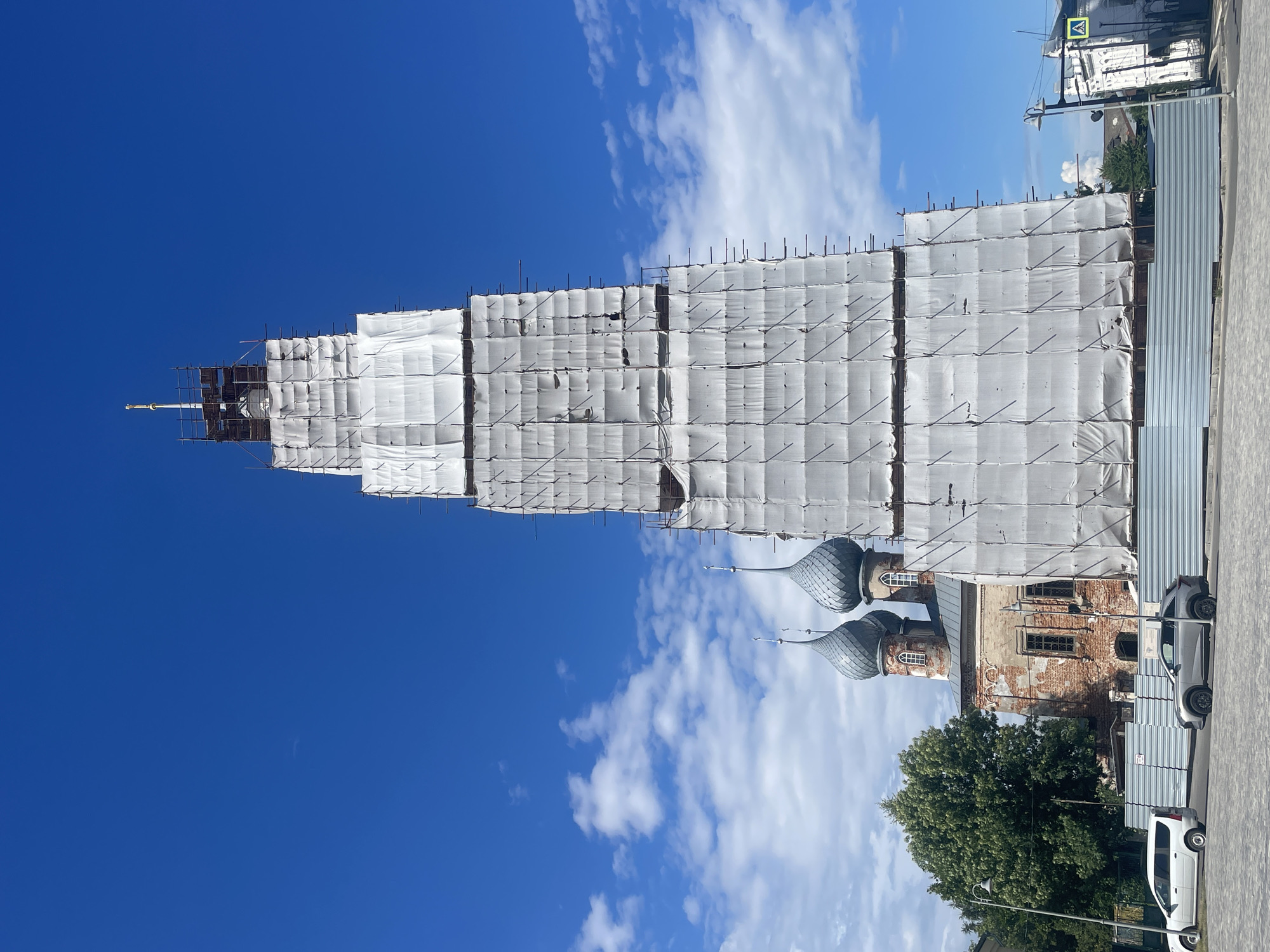 This is an outdoor, eye-level, medium-shot photograph of the Bell Tower of St. Nicholas Church in Yuryevets, Russia, under renovation. The bell tower, a tall, tiered structure, is largely covered by white tarpaulin, obscuring its architectural details. Scaffolding is visible around the tower. To the left of the bell tower are the church's main building and two of its onion domes, also in a state of disrepair, with exposed red brickwork. The sky is a vibrant blue with scattered white clouds. In the foreground, a gray car and a white minivan are parked on a paved street. A metal fence runs along the construction site. The overall impression is one of a historic building undergoing restoration against a clear blue sky.
