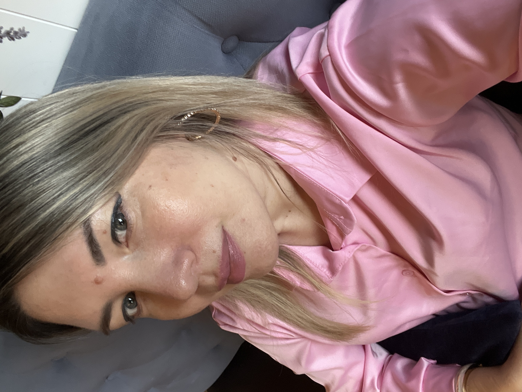 A woman with blonde hair and light skin is looking at the camera. She is wearing a pink satin shirt. She has on hoop earrings. Her lips are painted pink. The woman appears to be sitting on a gray sofa. The background shows a light-colored wall with some greenery and purple flowers. There are no elements in the image that indicate the location is the city of Sochi, Russia.