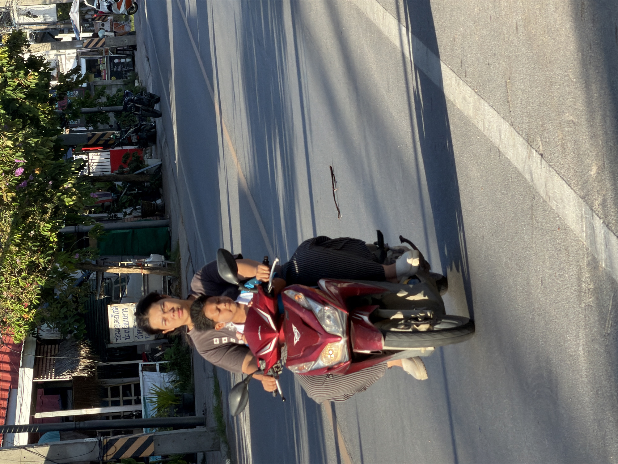 In the city of Ko Samui District, Thailand, a person is riding a red Honda scooter on an asphalt road. The rider, a woman with dark hair pulled back, is wearing a dark grey t-shirt and striped pants. A young boy with dark hair, wearing a white shirt, is seated in front of her. They are both wearing casual shoes. The scooter has a prominent front fairing with headlights. The scene is illuminated by sunlight, casting shadows on the road. In the background, there are buildings with red roofs, trees with green foliage and some pink flowers, and power lines. A few other motorcycles are visible parked in the background. The road appears to be a typical street in a commercial or residential area, with storefronts and pedestrian activity suggested by the presence of stalls.