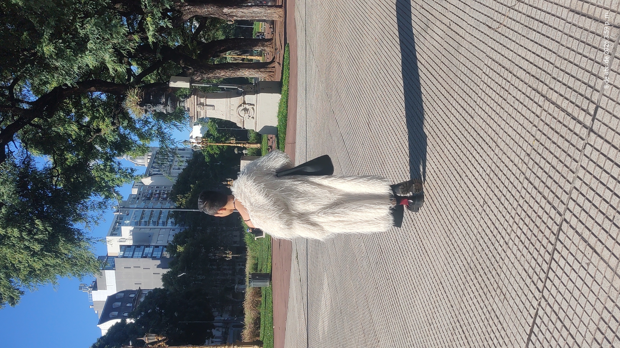 A full shot captures a person in a long, white, furry coat walking on a pedestrian walkway made of interlocking concrete pavers. The walkway is adjacent to a park area, which contains trees, lawn, and various monuments. The person wears dark shoes and black clothing below the coat.
Beyond the park area, buildings of Buenos Aires are visible against a clear blue sky. The large trees lining the park provide ample shade. The time stamp "8 de abr. de 2026 3:59 p. m." can be seen in the upper right corner of the image.