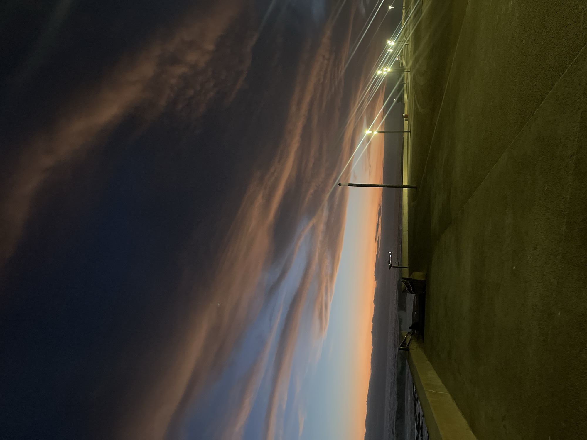 A nighttime scene in Arica, Chile. The sky is a gradient of deep blue and orange, with streaks of clouds. A line of streetlights illuminates a concrete walkway that runs along the edge of the coast. The ocean is visible in the distance. There are benches and possibly trash cans on the walkway. No people are clearly visible.