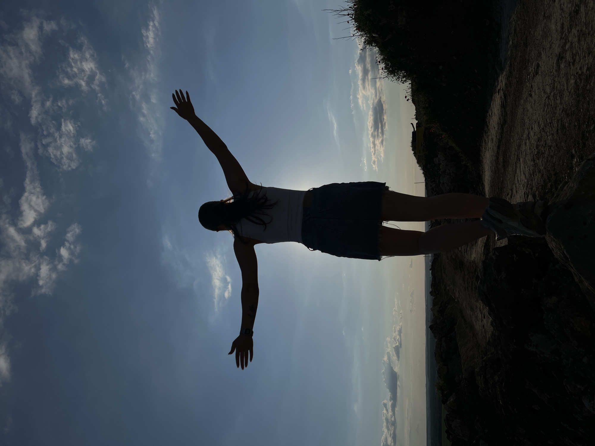 A silhouetted person stands on a rocky outcrop, arms outstretched towards a bright sky with scattered clouds. The person is viewed from behind, wearing a white tank top and a denim skirt. Their hair is pulled back in a ponytail. The sun is positioned behind and slightly to the right of the person, creating a lens flare effect. The ground beneath them is uneven and appears to be a dirt path with rocks. In the distance, a vast landscape stretches out, with fields and some distant structures visible under the diffused sunlight. The overall impression is one of freedom and openness against a backdrop of a wide sky and expansive terrain. The location is identified as Khomivka, Ukraine, suggesting an outdoor setting in a rural or semi-rural area.