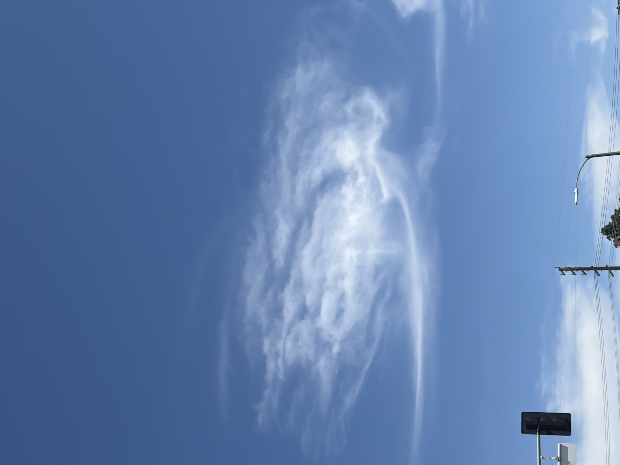 The image shows a vertical shot of a blue sky with cirrus clouds. On the right side of the frame, there is a street scene with utility poles and lines. A street light and a rectangular traffic signal are also visible. The location is likely in the city of Los Angeles, United States of America, based on the urban infrastructure and the type of utility poles.