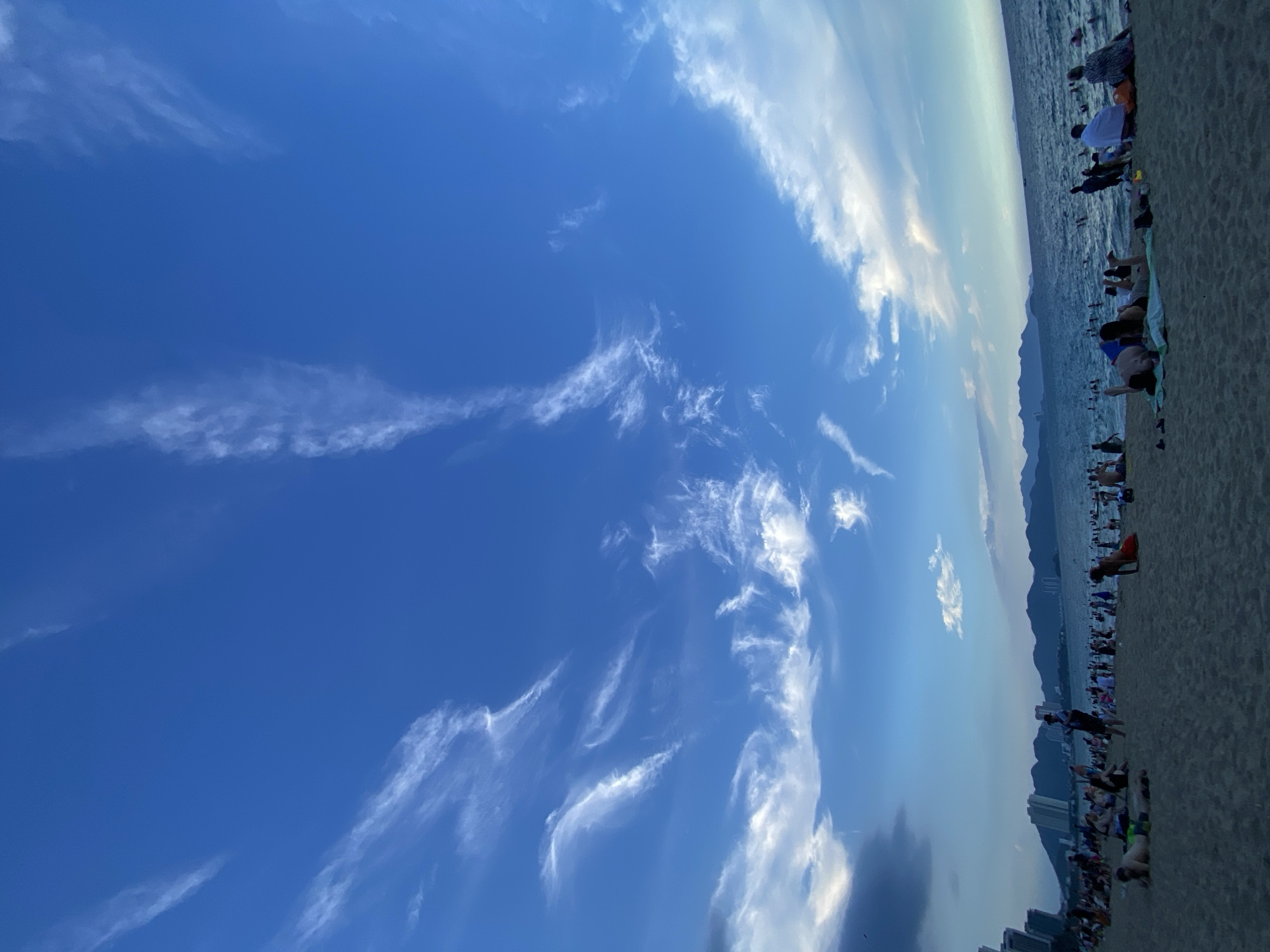 The image depicts a crowded beach scene in Nha Trang Ward, Vietnam, under a vibrant blue sky with wispy white clouds. The foreground is dominated by a sandy beach populated with numerous individuals, many of whom are lying down or sitting. Some are dressed in swimwear, while others wear casual clothing. A section of the sea is visible in the midground, with people swimming and wading. In the background, a city skyline with tall buildings lines the coast, and a mountain range stretches across the horizon. The overall atmosphere appears to be one of leisure and recreation.