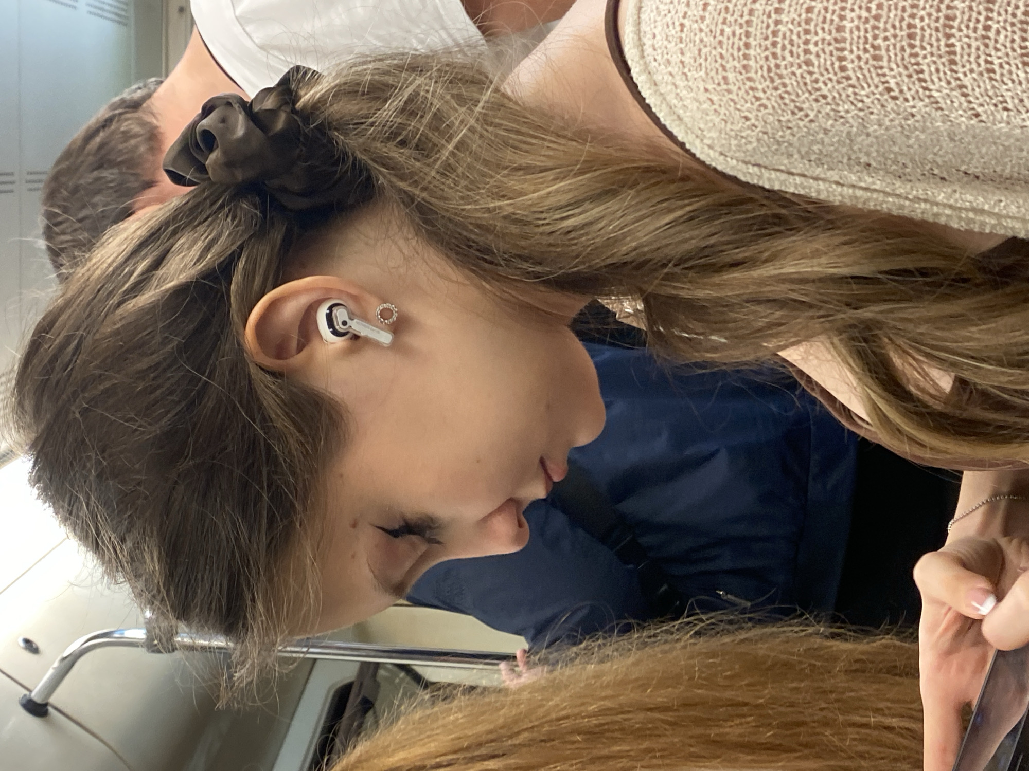 A young woman with long, wavy brown hair tied in a ponytail with a dark brown scrunchie is seen in profile, looking down and to her left. She has fair skin, her eyes are closed, and she is wearing white wireless earbuds with silver accents and a small hoop earring in her left ear. Her right arm is visible, holding a dark object, likely a smartphone, and she has a fine silver bracelet on her wrist. She is wearing a cream-colored open-knit cardigan over a brown garment. In the background, a man with short brown hair is partially visible, wearing a white t-shirt. To the left of the young woman, another person with long, light brown hair is visible. The environment appears to be a public transportation setting, possibly a subway car, indicated by a silver pole and handrail structure visible in the upper left corner. The lighting is even and appears to be artificial. The overall impression is of a candid moment captured within a bustling urban environment, possibly during a commute in Moscow, Russia.