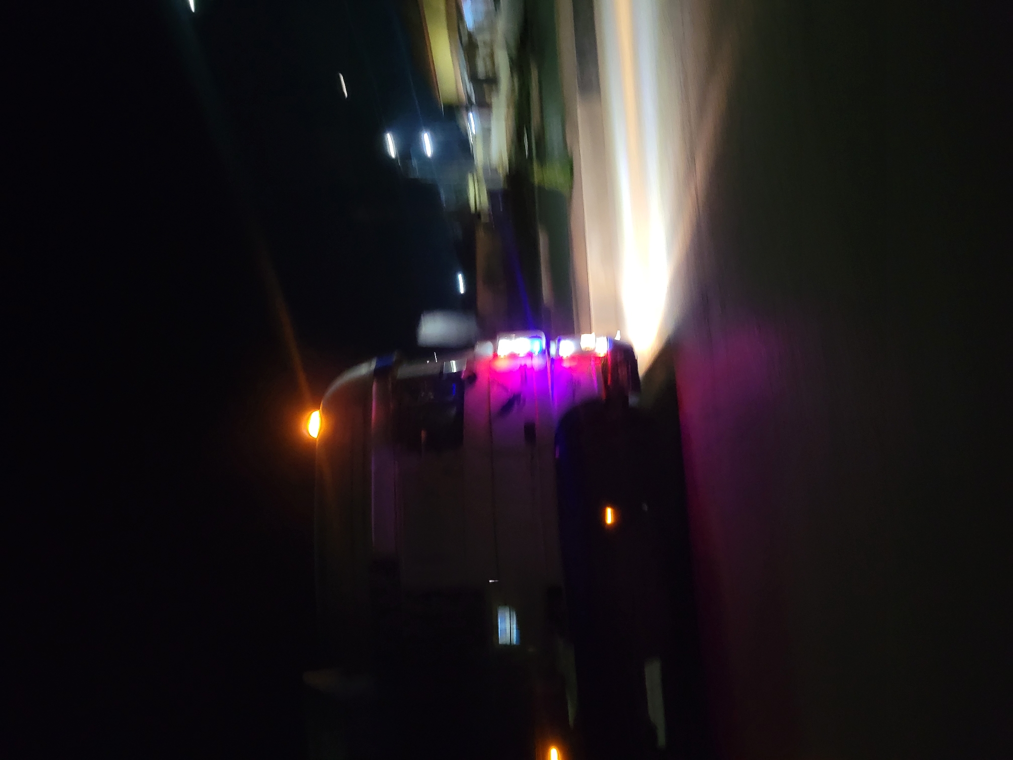 The image shows a vehicle with emergency lights, possibly an ambulance or fire truck, parked on a street at night. The location appears to be urban, with streetlights and buildings visible in the background. The vehicle has red, blue, and white flashing lights on its roof. The image is slightly blurry, suggesting movement during the capture.