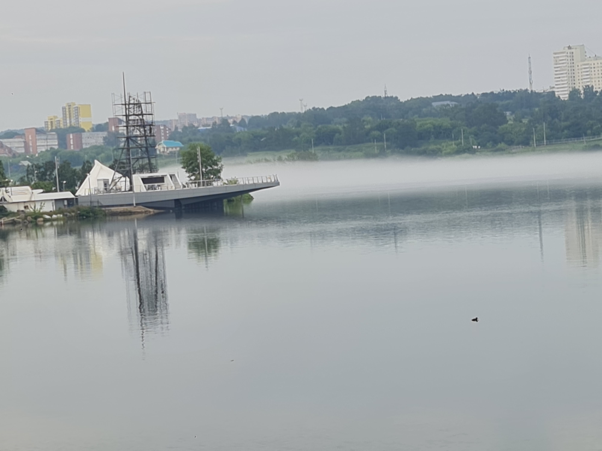 In an outdoor, waterfront scene, a tall, dark, skeletal structure resembling a ship's mast and rigging rises from a partially constructed or renovated boat-shaped platform on the water. The platform, a mix of white and grey, has a modern design and appears to be undergoing some form of construction or refurbishment. Adjacent to it, a white, tent-like structure is visible.
The body of water is calm, reflecting the overcast sky and the structures on the shore. A thin layer of fog hovers low over the water, partially obscuring the background. The background reveals a city skyline with several tall buildings and residential blocks, interspersed with trees and green hills. The overall atmosphere is one of a slightly hazy, quiet morning. On the water in the middle ground, a small, dark object, possibly a duck or a piece of debris, is visible. The reflection of the skeletal mast and parts of the platform are clearly visible in the water.