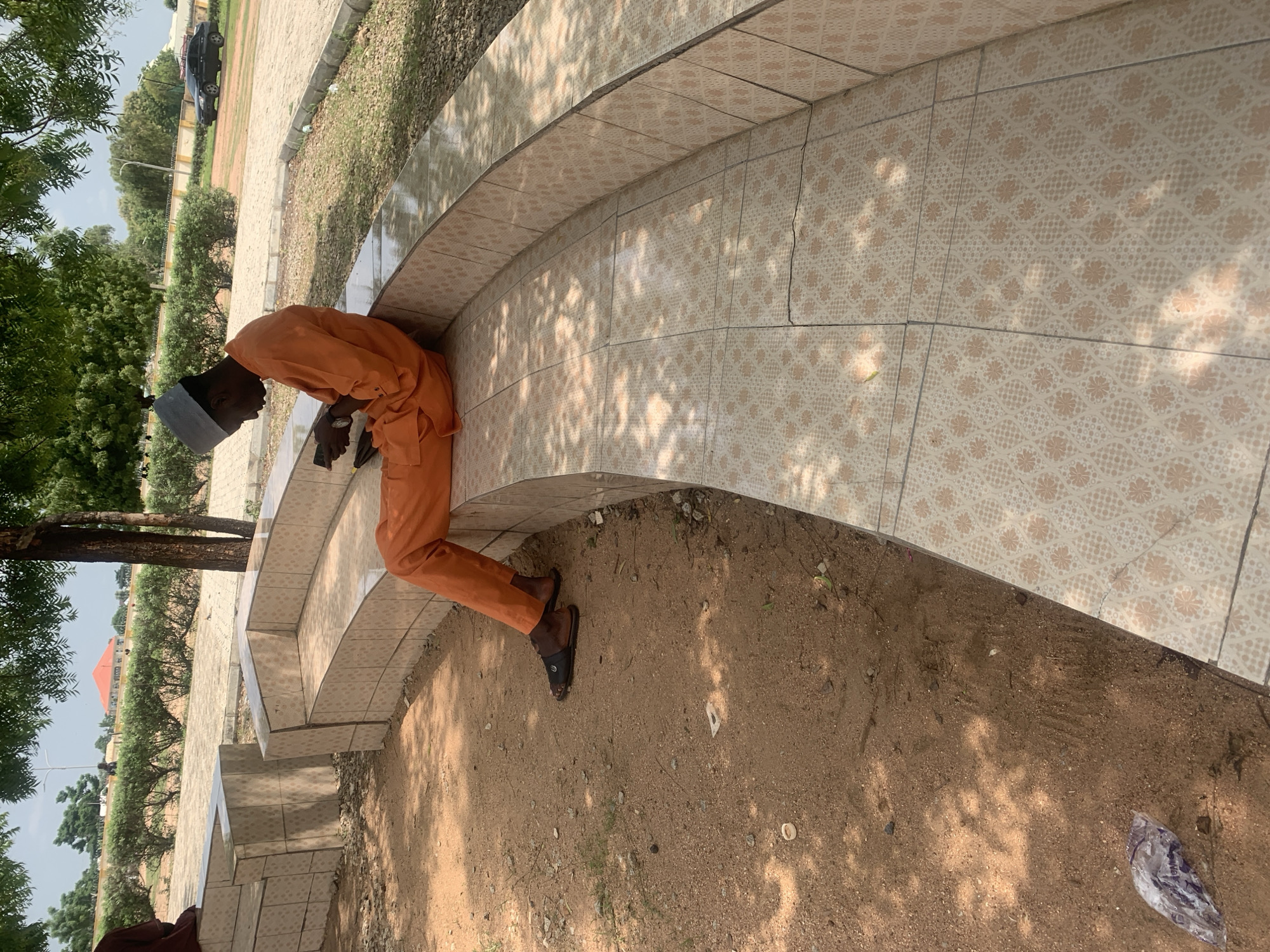 A young man wearing an orange garment and a grey cap is sitting on a curved, tiled bench in an outdoor area, looking down at his phone. The bench is made of light-colored tiles with a floral pattern. The ground around the bench is sandy with some scattered debris, including a plastic bag. In the background, there are green bushes, trees, a paved area, and a building with an orange roof. A black car is parked in the distance. The scene appears to be in Jalingo, Nigeria, possibly a public park or rest area.