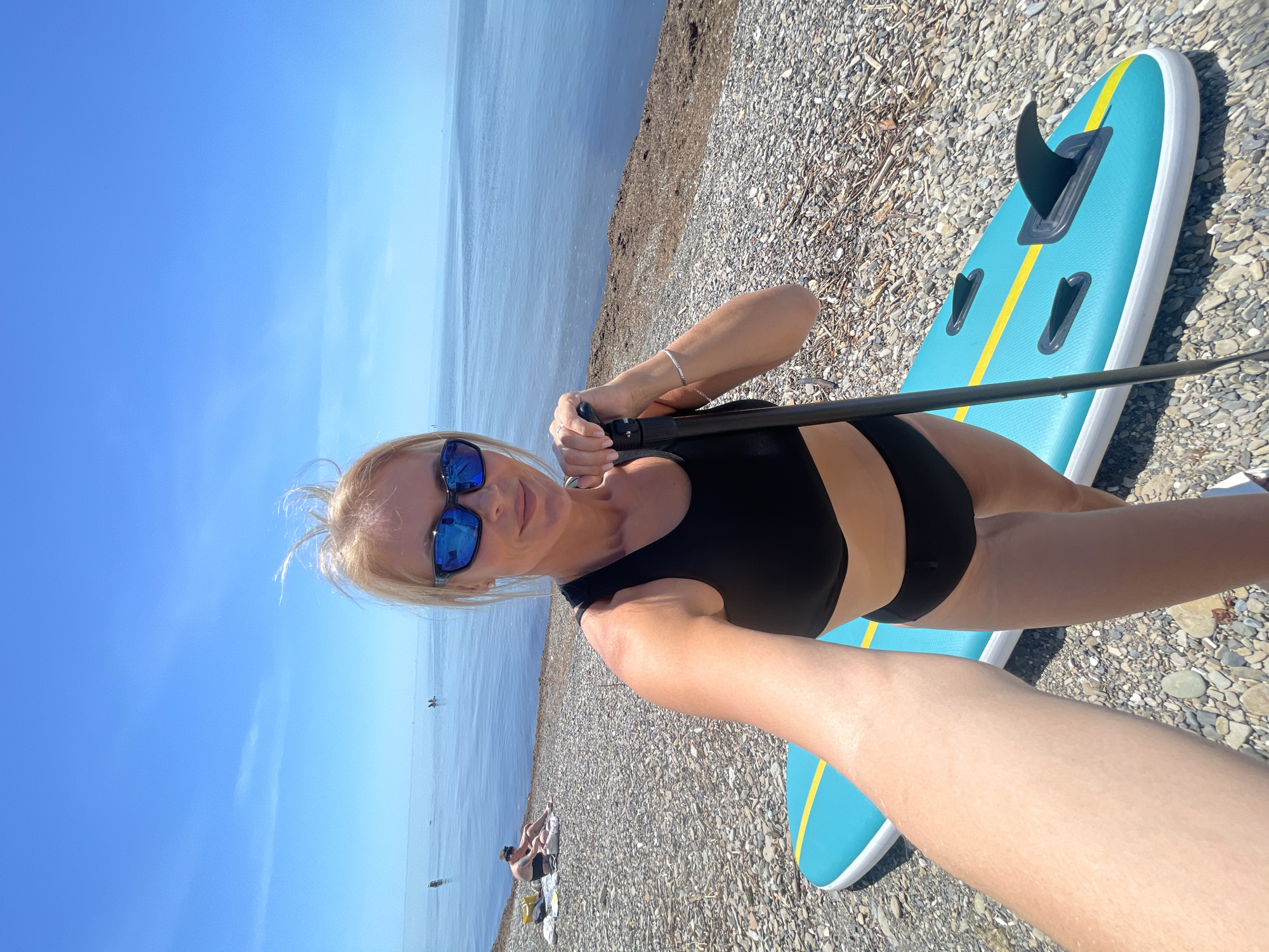 A blonde woman is captured in a selfie on a shingle beach in Sochi, Russia, facing the camera with a slight smile. She wears blue mirrored sunglasses, a black sports bra or bikini top, and black bottoms. Her left hand holds the handle of a paddle, suggesting she is preparing for or has just completed stand-up paddleboarding. Beside her, a blue and yellow stand-up paddleboard with visible fins rests on the pebbly shore.
The scene depicts a bright, sunny day with a clear blue sky. The calm, blue sea stretches out to the horizon, dotted with a few distant boats or other watercraft. Further along the shingle beach, another person can be seen relaxing in the distance. The strong sunlight indicates it is likely midday or early afternoon. No visible text is present in the image. The overall setting is one of leisure and outdoor water activity.