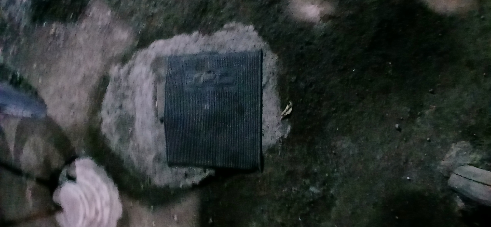 A dark rectangular doormat with a ribbed surface is positioned on a patch of exposed soil. The doormat appears to be black or a very dark color. Surrounding the doormat is a mixture of soil and grass. In the background, a light-colored, fluffy object is visible, possibly a piece of fabric or padding. The overall scene is outdoors, and the lighting suggests it may be dusk or nighttime. There is no indication of people or specific landmarks that would pinpoint a location within Demangan, Indonesia.