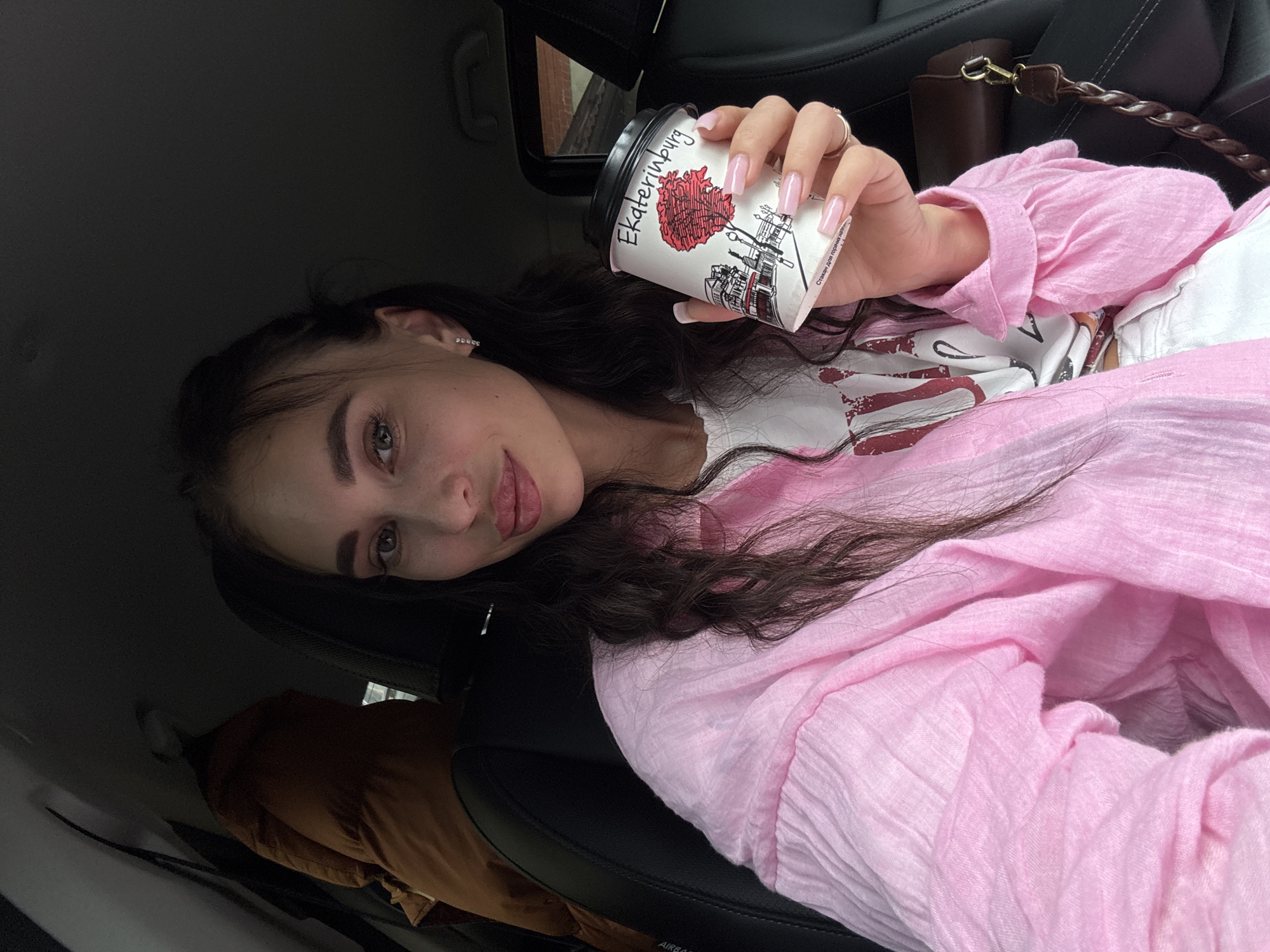 A young woman with dark, wavy hair and striking makeup is seated in the driver's seat of a car. She is wearing a pink, crinkled fabric shirt over a white t-shirt and holding a disposable coffee cup with a picture of a tree and buildings on it. The text on the cup reads "Ekaterinburg". Her fingernails are long and painted a light pink. She is looking at the camera with full lips slightly parted. The car's interior is visible, including a black leather seat and a tan jacket hanging on the back of the passenger seat. The background suggests an outdoor, possibly urban, environment.