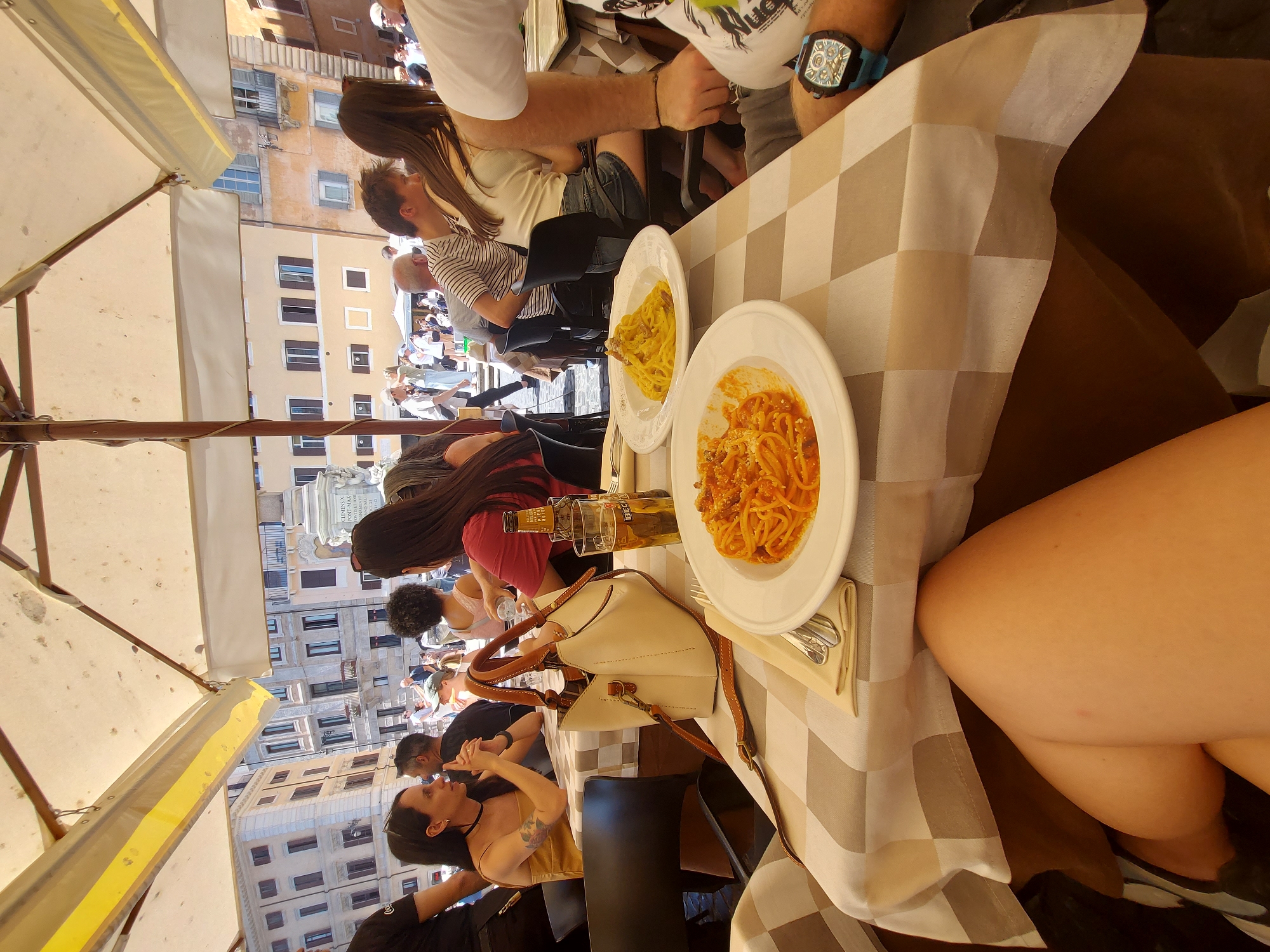 The image depicts a group of people dining at an outdoor restaurant in Rome, Italy, potentially in the vicinity of Piazza Navona. Several individuals are seated at tables covered with checkered tablecloths under the shade of sun umbrellas. Plates of pasta, possibly spaghetti, are visible on the tables. The background shows buildings and a fountain, indicative of a historical city square. People are passing by, suggesting a busy public space. A beige handbag rests on the table near a beer bottle. Some diners are engaged in conversation.